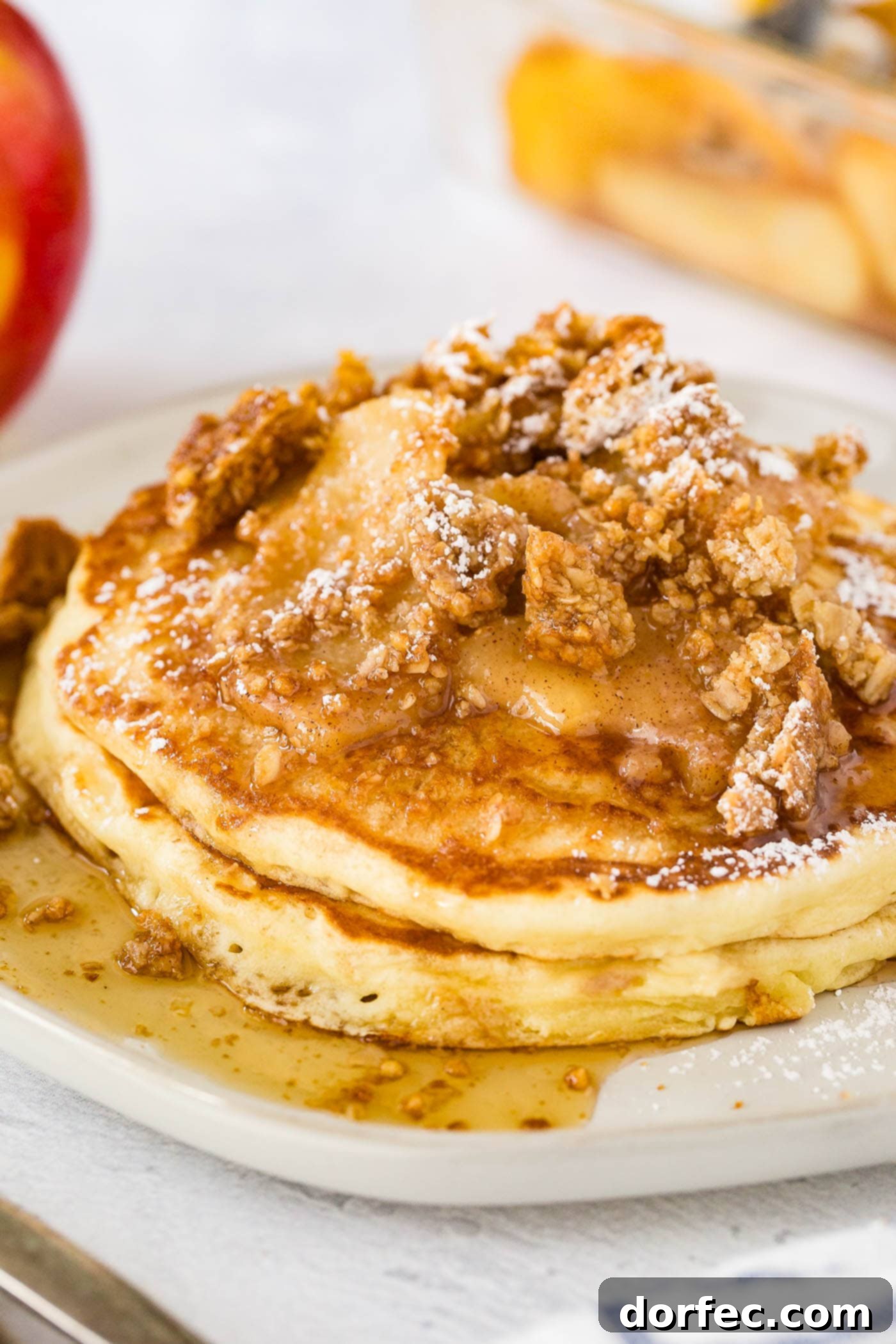 A beautifully composed plate features a towering stack of golden brown pancakes, generously adorned with tender, caramelized cinnamon apples, a sprinkle of crisp granola, and a delicate dusting of powdered sugar. A shimmering ribbon of maple syrup cascades over the top, with a side of perfectly cooked bacon adding a savory contrast.