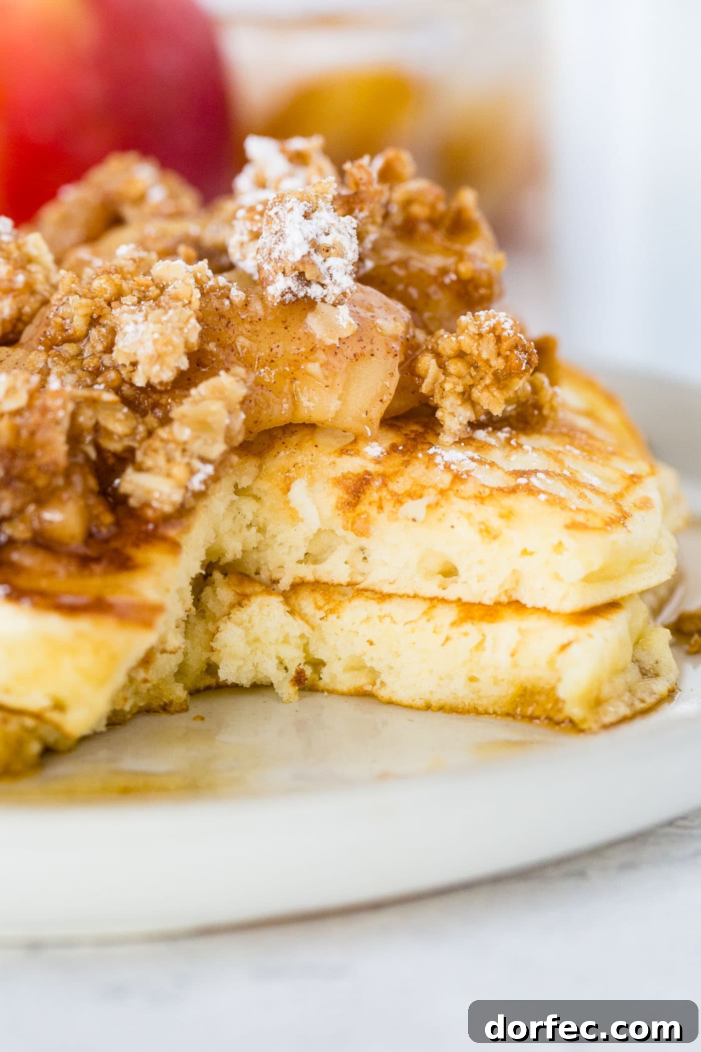 A close-up view of a vibrant breakfast plate featuring a stack of golden pancakes, richly topped with cinnamon-roasted apples, a sprinkle of granola, and powdered sugar. A stream of maple syrup adds a glossy finish, accompanied by a side of crispy bacon.