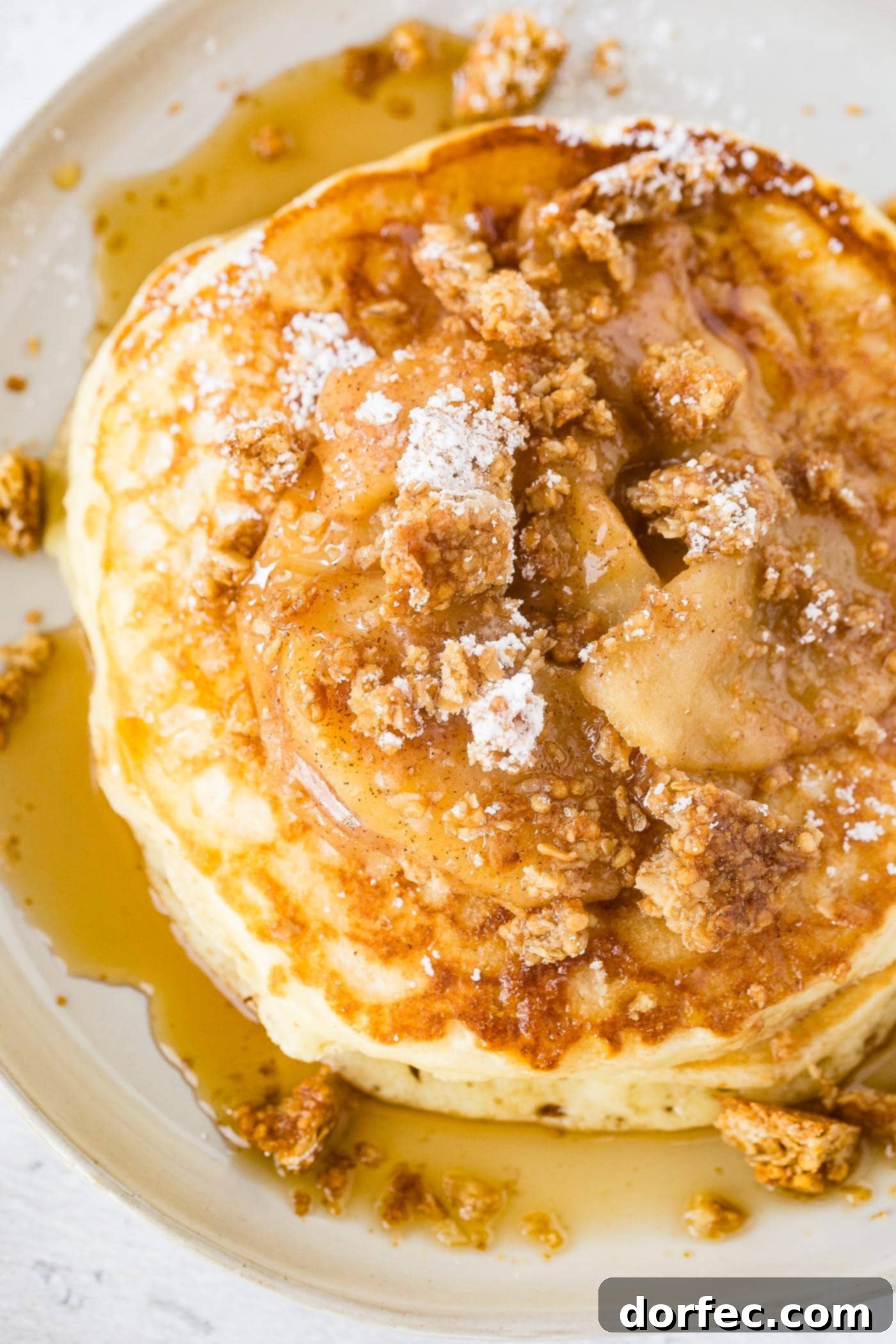 A stacked plate of golden pancakes, lavishly topped with cinnamon-roasted apple slices, a generous scattering of granola, and a delicate dusting of powdered sugar. A glossy stream of maple syrup cascades over the top, with a side of crispy bacon.