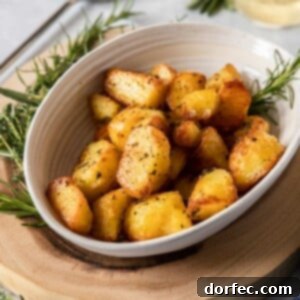 A cream-colored bowl filled with herby roasted potatoes, surrounded by fresh herbs. It sits on a wooden board