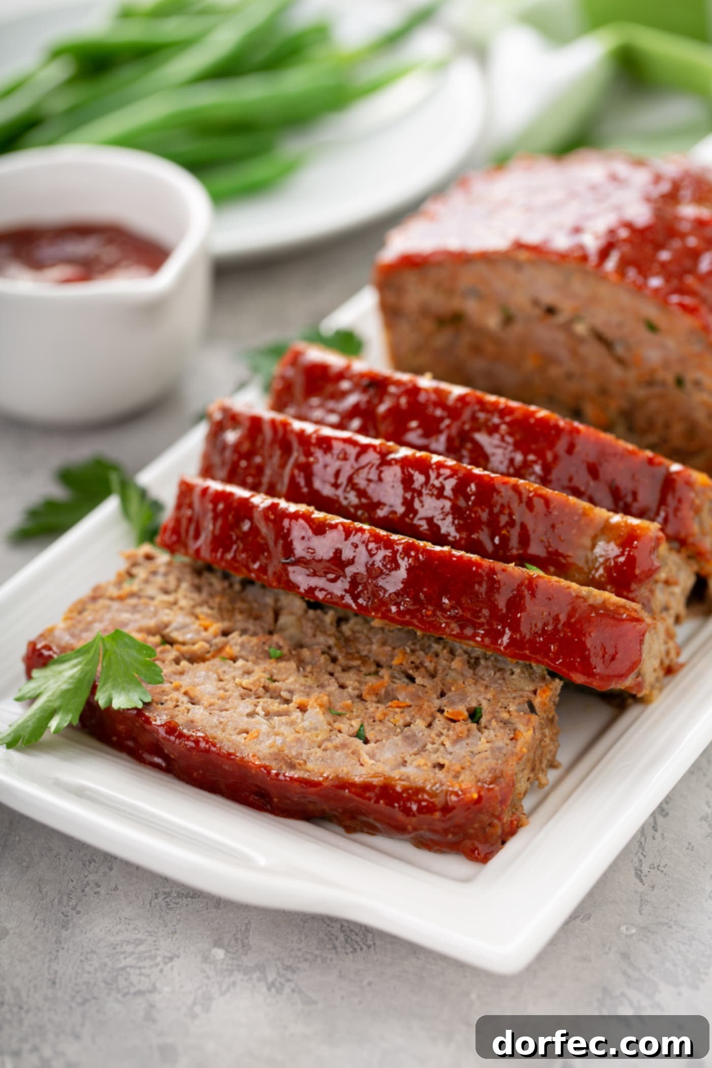 A close-up of a meatloaf on a serving tray, half-sliced to reveal its juicy interior, with a small bowl of savory glaze nearby and green beans blurred in the background.