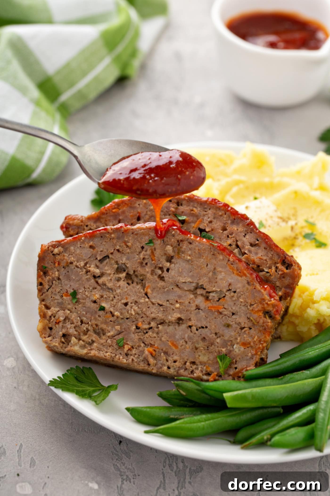A delectable dinner plate featuring mashed potatoes, green beans, and two slices of classic meatloaf, with a rich red glaze being artfully drizzled over the top.