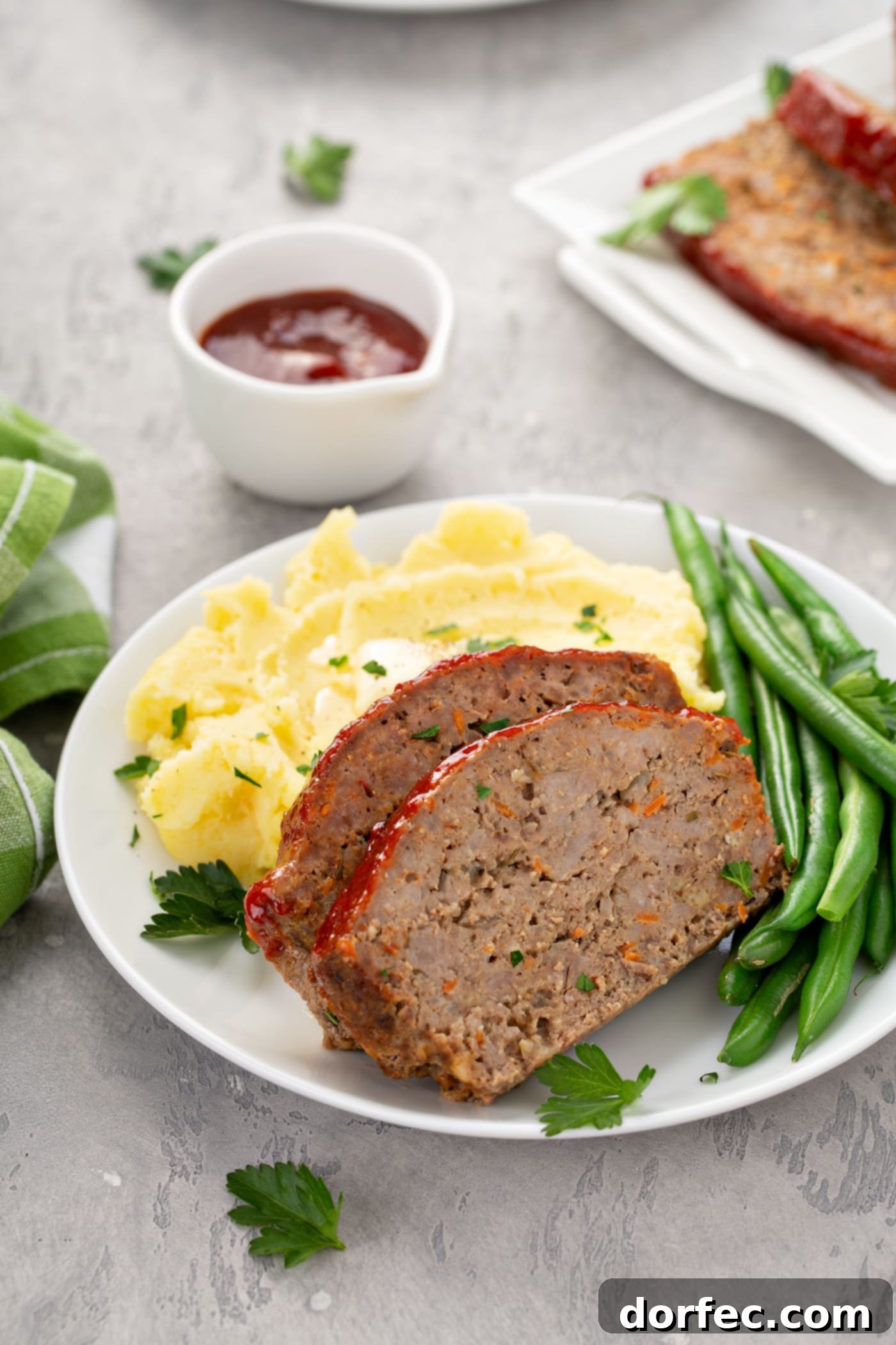 A white dinner plate holding two tender slices of meatloaf, accompanied by a side of fresh green beans and creamy mashed potatoes, with a small container of glaze in the background.