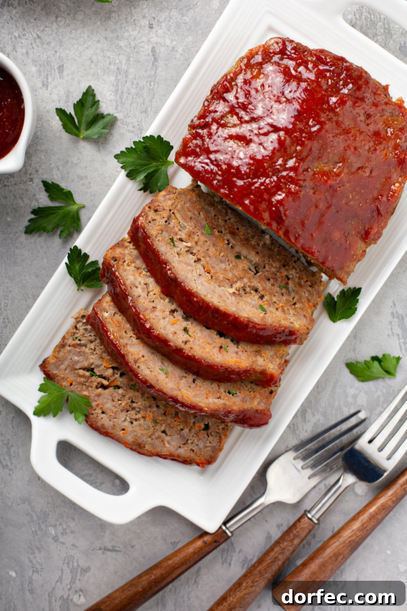 A rustic serving tray showcasing a partially sliced meatloaf, garnished with fresh parsley. Two forks rest beside the tray, and a bowl of rich glaze waits in the background.