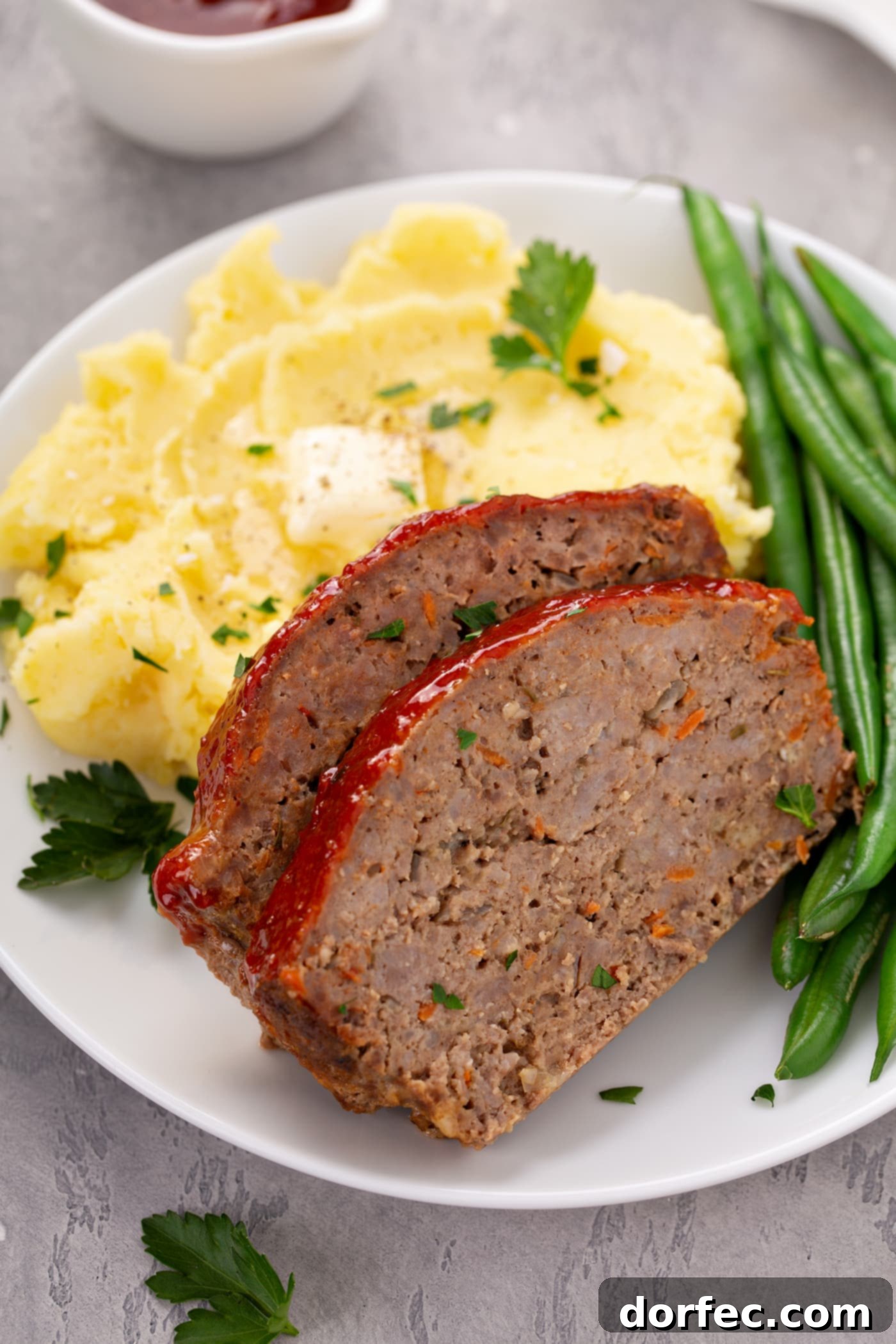 A beautifully plated dinner with two slices of meatloaf, a generous serving of mashed potatoes topped with melting butter and fresh parsley, and crisp green beans.