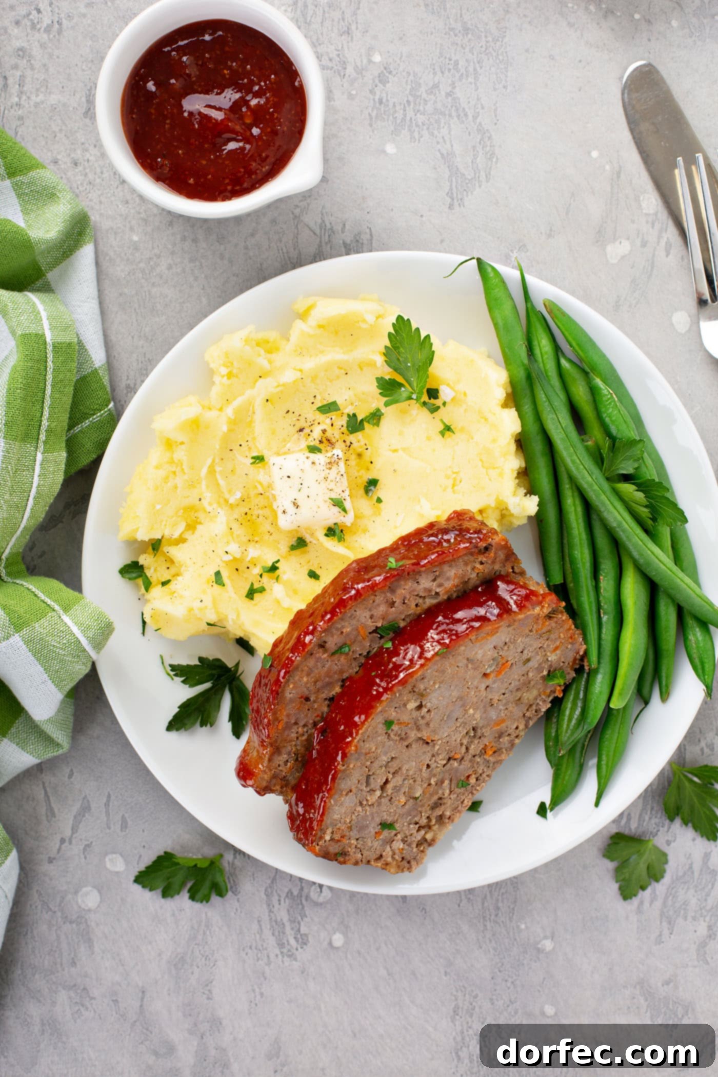 A complete dinner plate featuring two slices of meatloaf, mashed potatoes with butter and parsley, and green beans. A green plaid napkin and a container of red glaze complement the setting.
