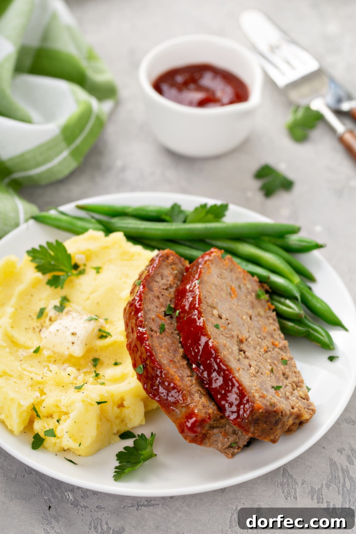 A comforting dinner plate with two slices of meatloaf, green beans, and creamy mashed potatoes topped with melting butter. A green plaid napkin and a container of red glaze are in the background.