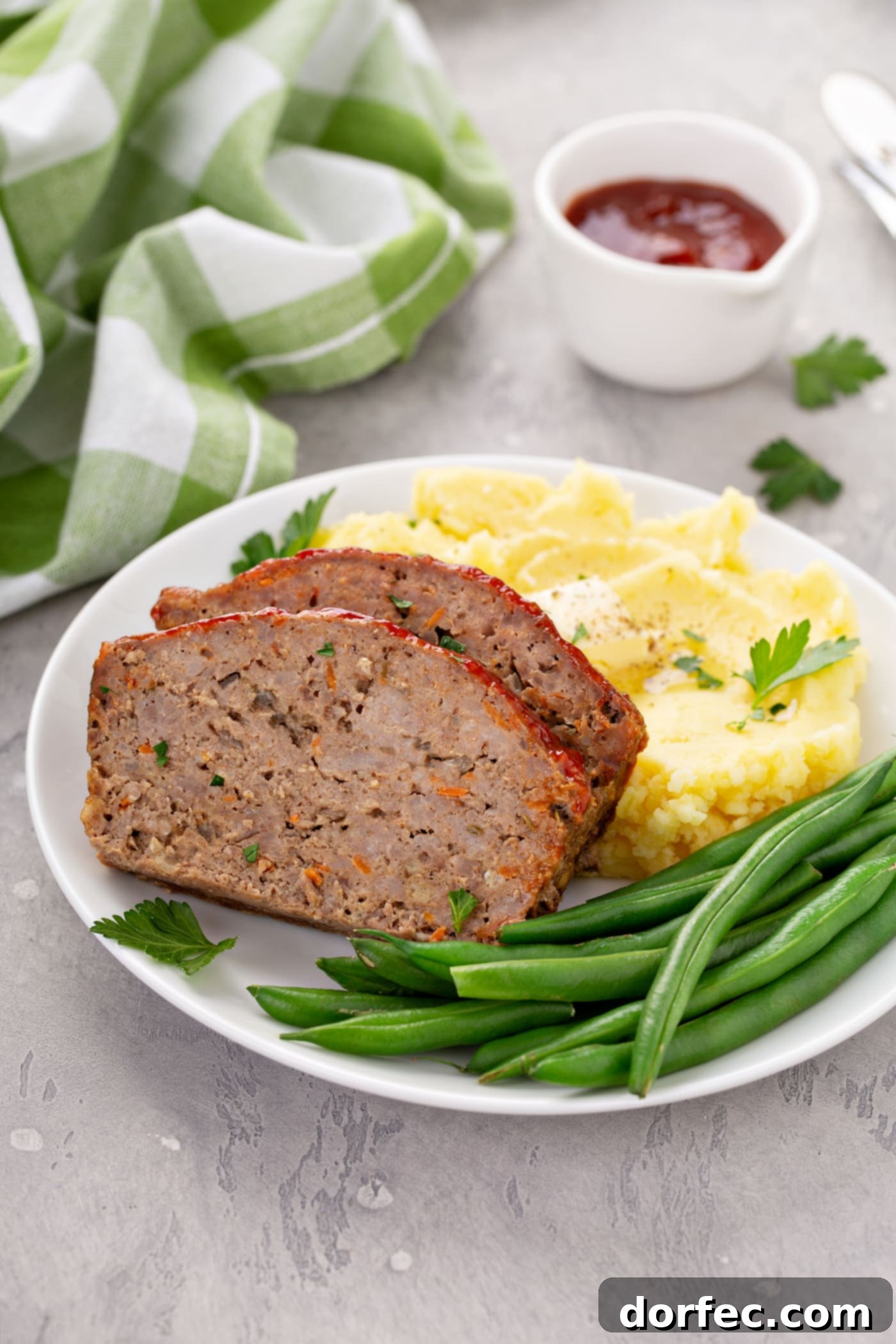 A hearty and appealing dinner plate showcasing two slices of meatloaf, green beans, and fluffy mashed potatoes adorned with butter and parsley. A green plaid napkin and a container of red glaze complete the inviting spread.