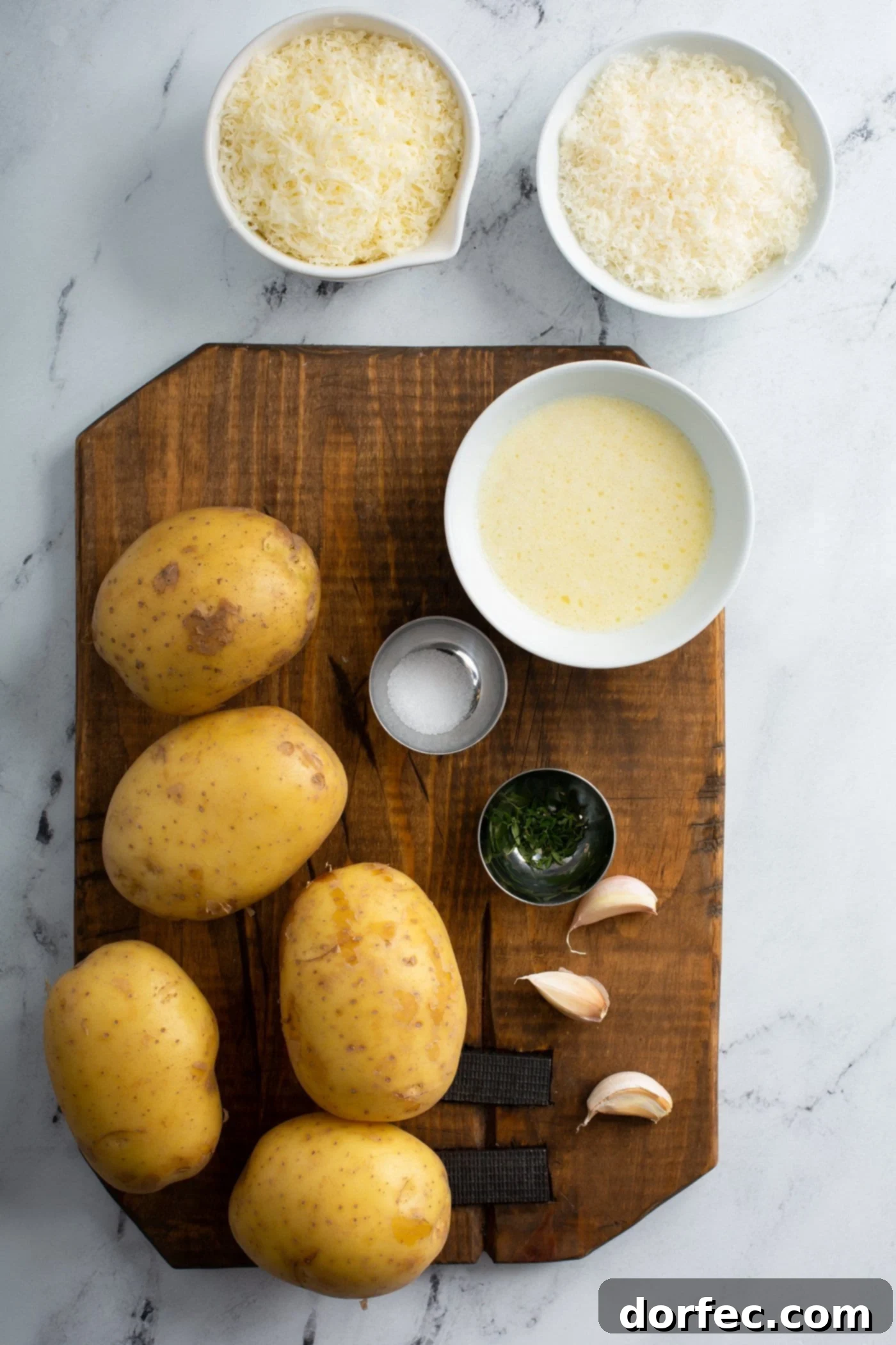 A wooden cutting board with four potatoes, three cloves of garlic, salt, pepper, melted butter, and cheese.