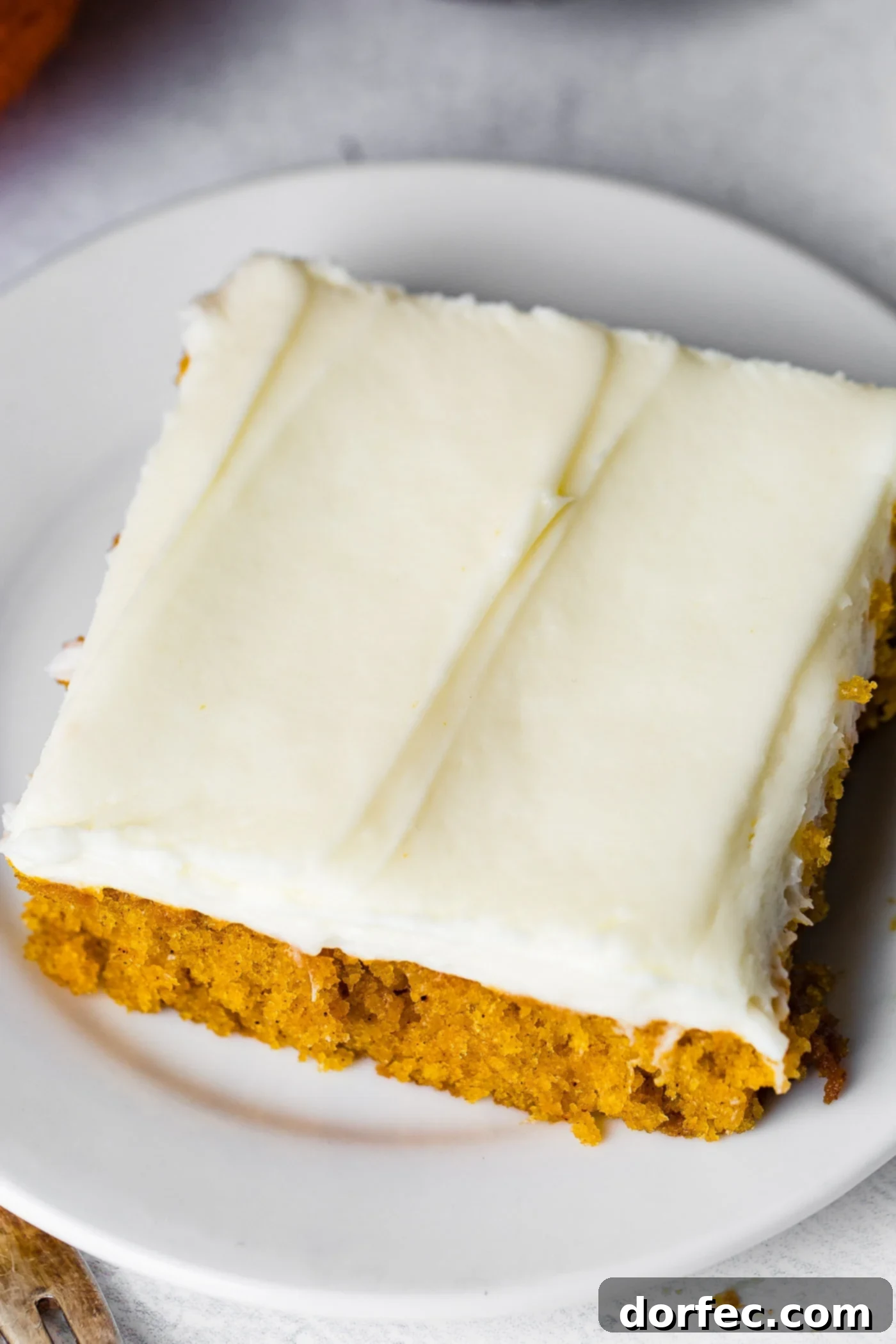A photo of a piece of pumpkin sheet cake with cream cheese frosting sitting on a white plate.