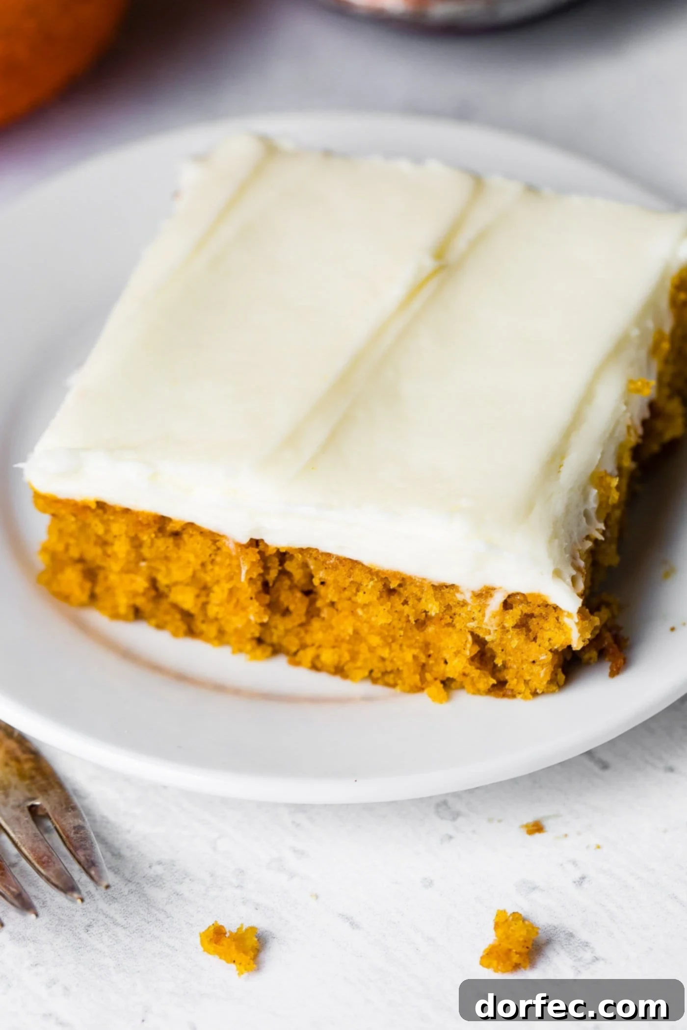 A photo of a piece of pumpkin sheet cake with cream cheese frosting sitting on a white plate.