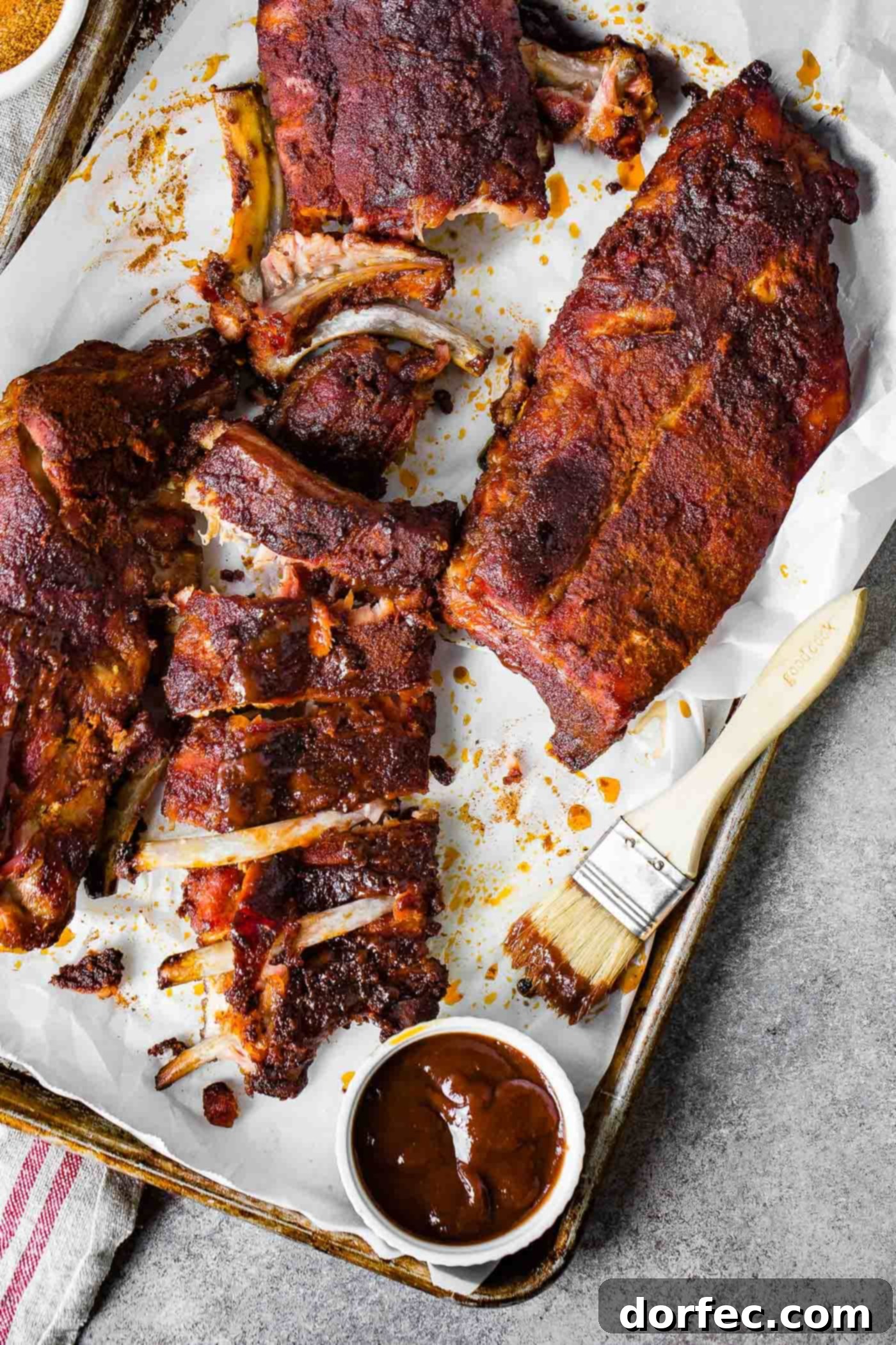 Traeger Smoked Ribs Three racks of exquisitely smoked ribs, two expertly sliced into serving portions and one presented whole, accompanied by a sauce brush and a bowl of delicious BBQ sauce.