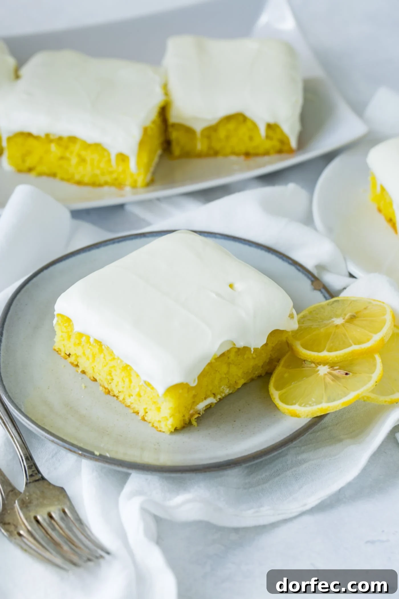 A vibrant slice of frosted lemon jello cake adorned with two delicate lemon slices, resting on an elegant serving plate. In the softly blurred background, another plate showcases more enticing cake slices, all nestled on crisp white linen napkins.