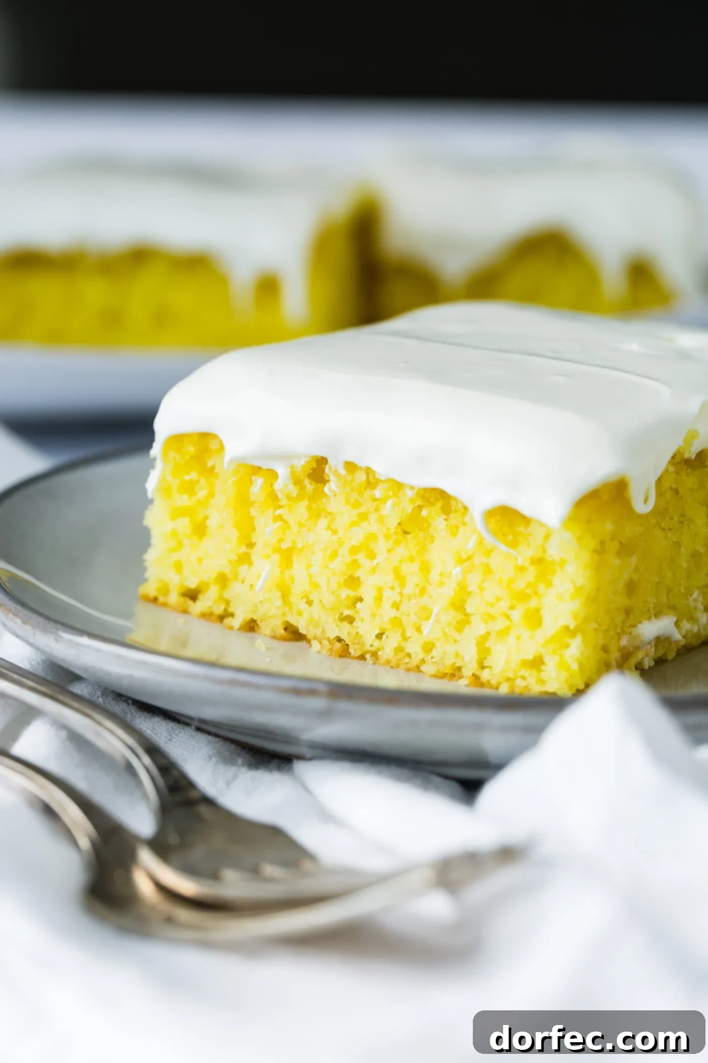 A tempting slice of frosted lemon jello cake graces a serving plate, with additional slices artfully arranged on another plate in the background. Two gleaming forks rest gracefully on a pristine white linen napkin beside the cake, inviting indulgence.