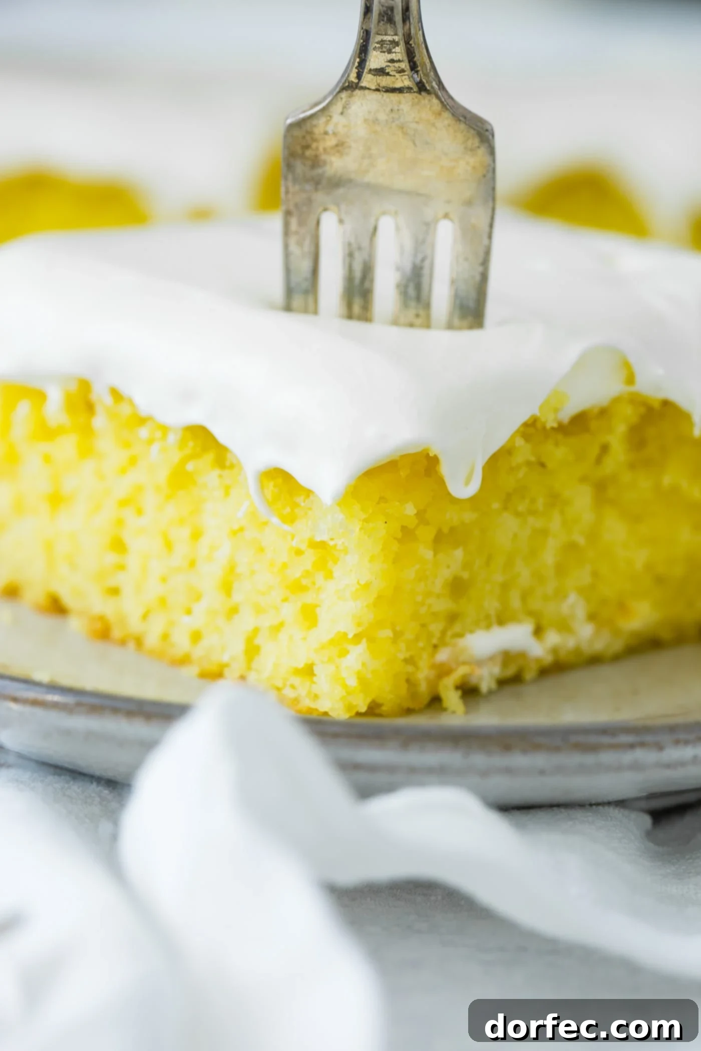 A delectable slice of frosted lemon jello cake, with a fork gently piercing its fluffy topping, rests on a pristine serving plate, artfully placed on a soft white linen napkin. #dessert #cake #lemon #frosting
