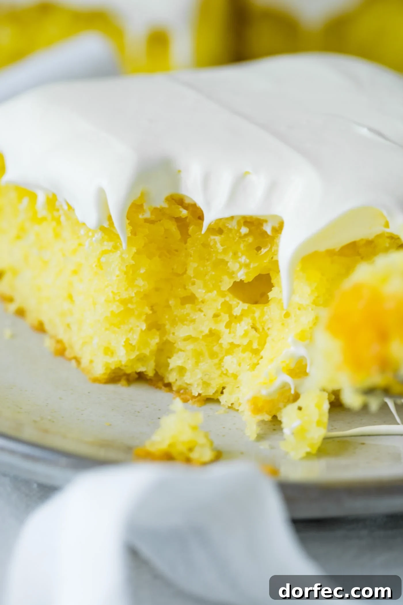 A bright yellow lemon jello cake slice with a cloud of fluffy white frosting on top. A bite has been delightfully taken from the corner, revealing the soft texture within.