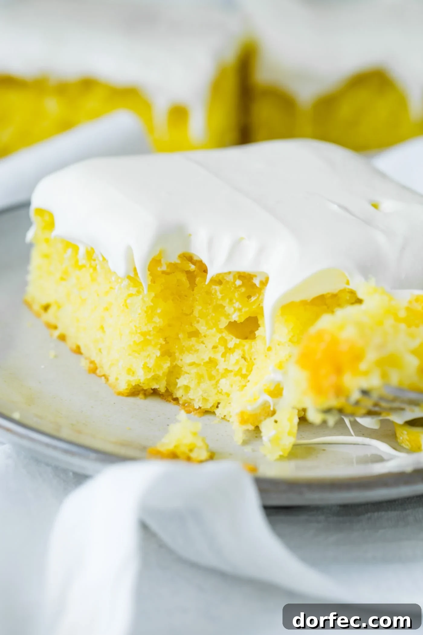 A delectable slice of vibrant yellow lemon jello cake, with a tempting bite taken from its corner. It's crowned with a cloud of fluffy white frosting and elegantly rests on a serving plate, which is in turn placed on a soft white linen napkin. #lemon #cake #dessert #frosting