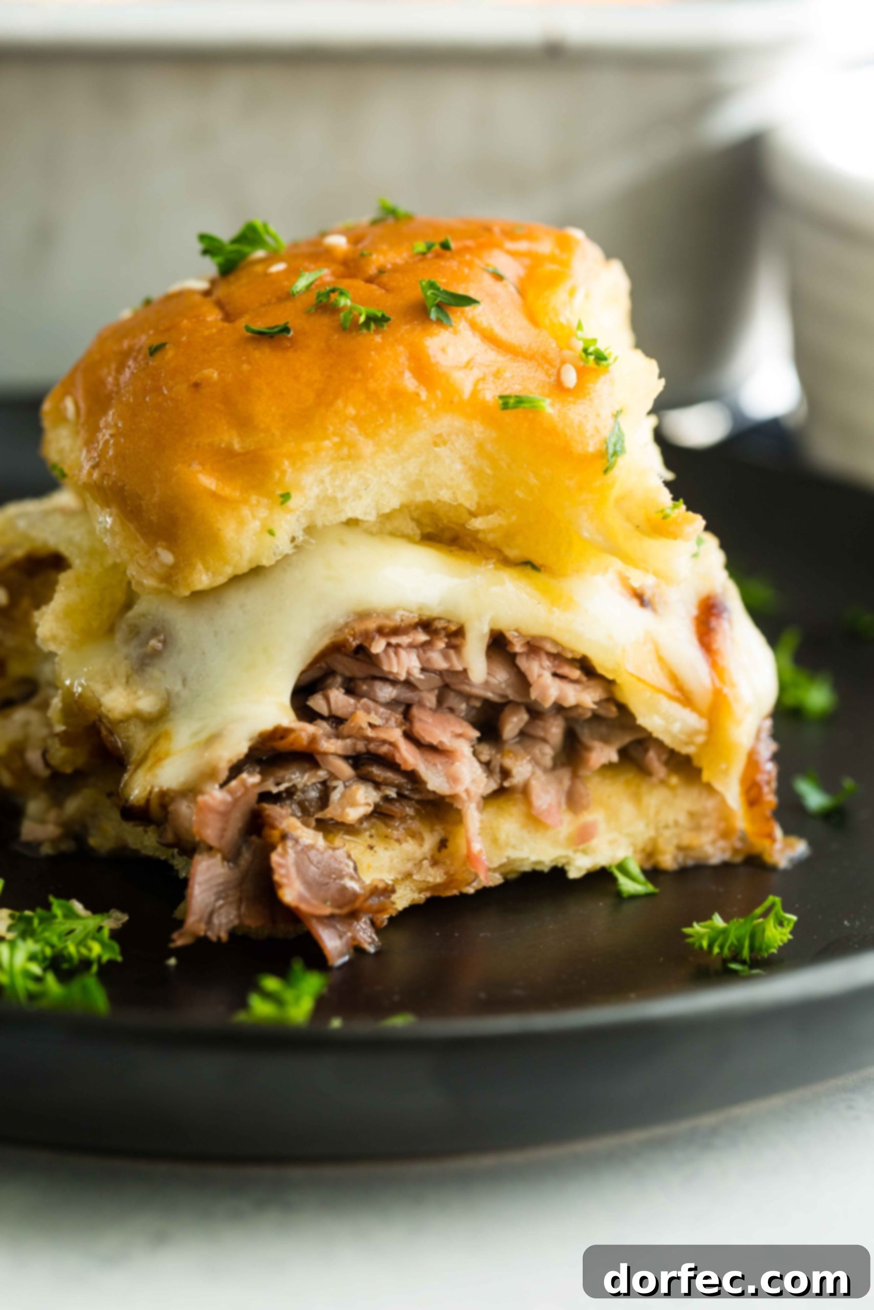 A photo of a single french dip slider on a black plate, showcasing its golden bun, melted cheese, and roast beef.
