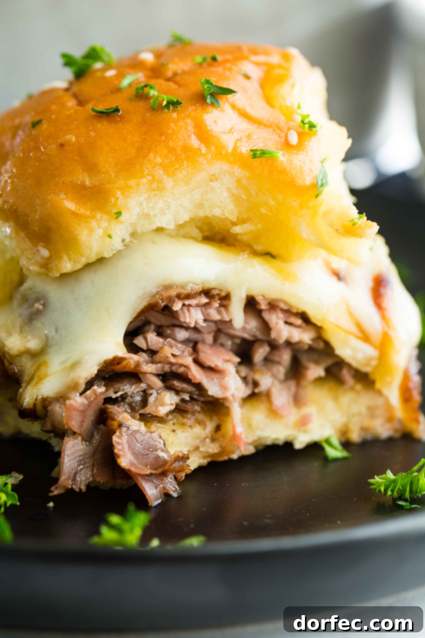 A photo of a single french dip slider on a black plate, showcasing its melted cheese and golden bun.