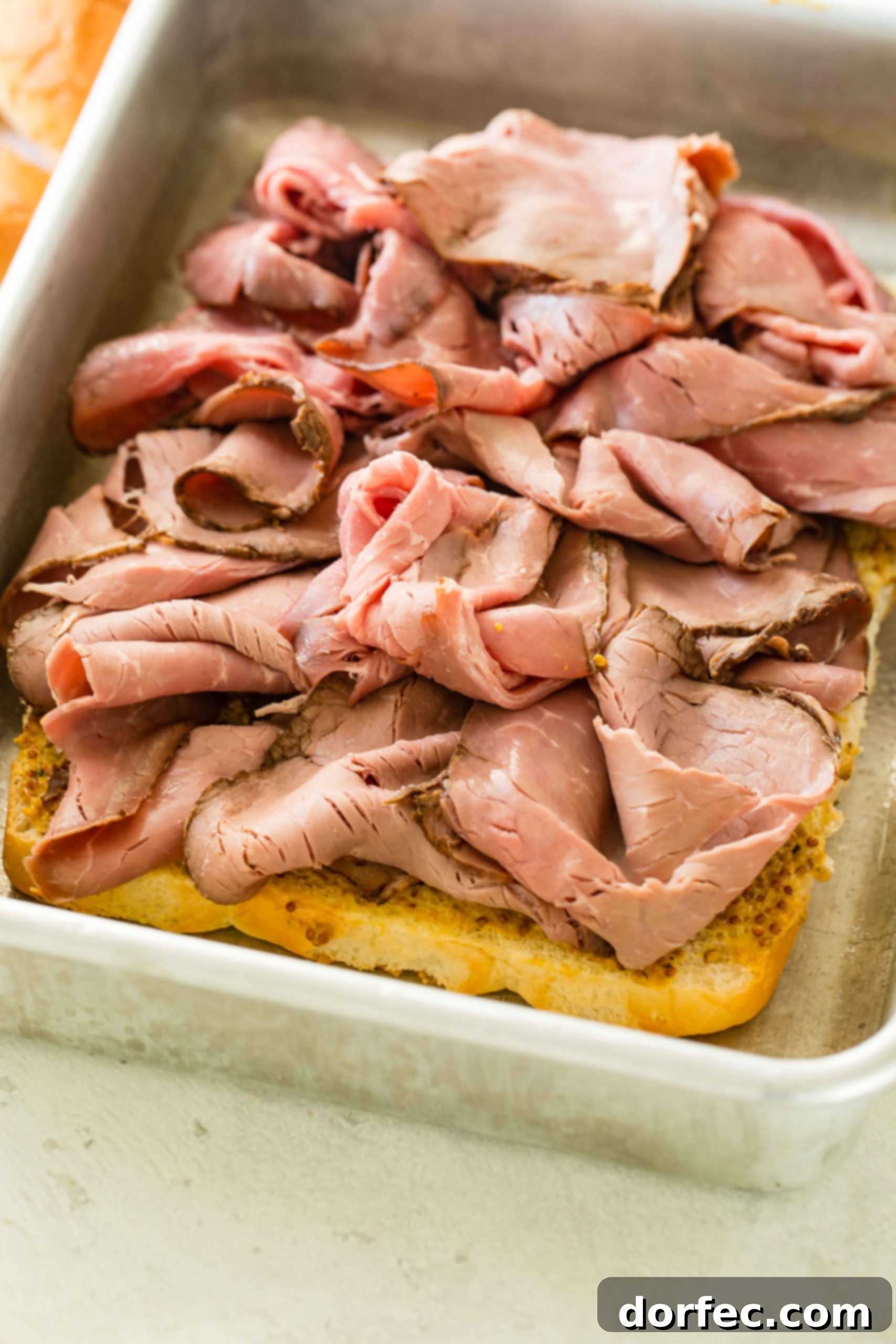 A photo of french dip sliders being constructed with a layer of roast beef added, covering the mustard buns.