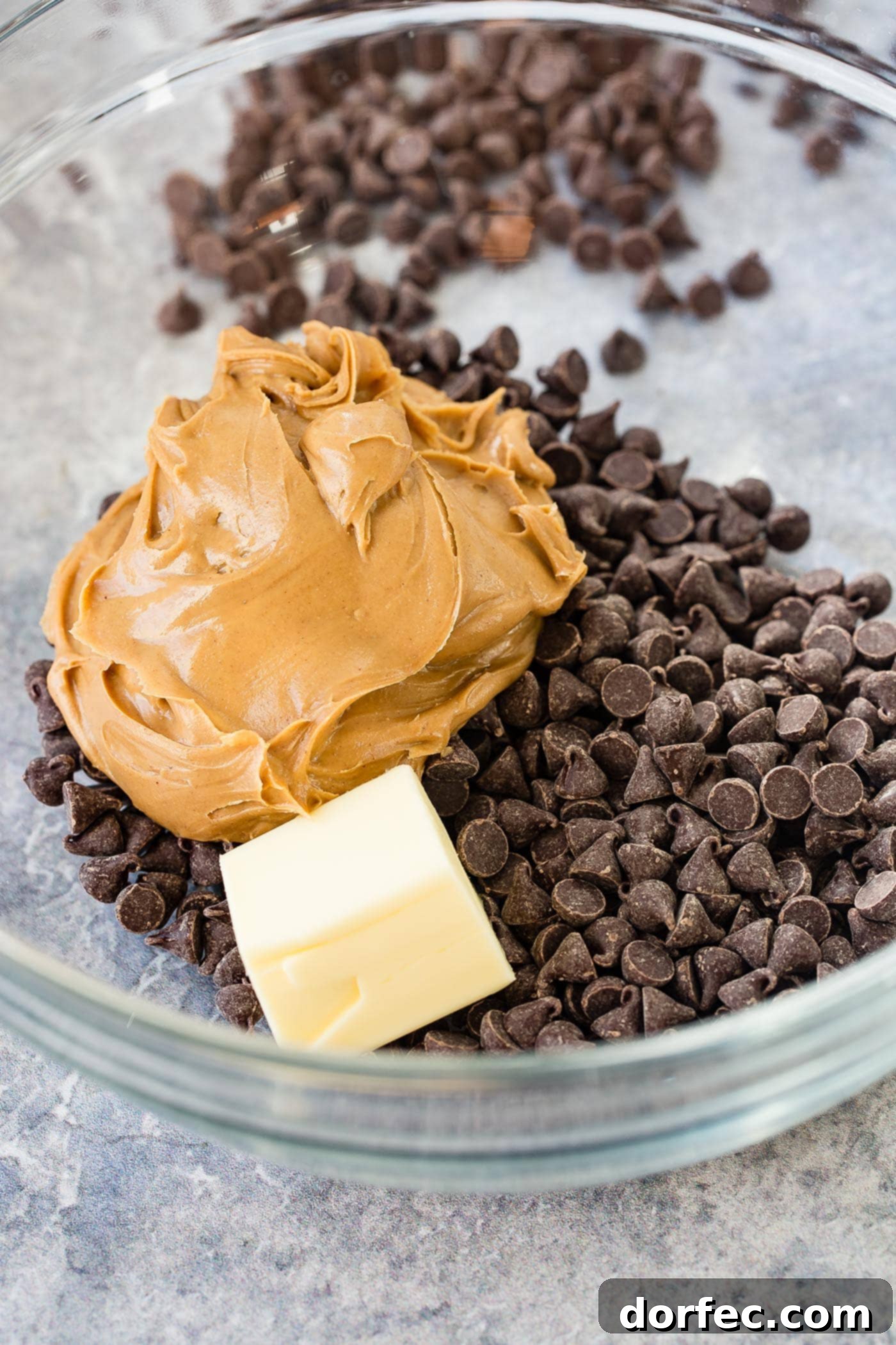 A glass bowl full of chocolate chips, a large dollop of peanut butter and a chunk of unmelted butter.