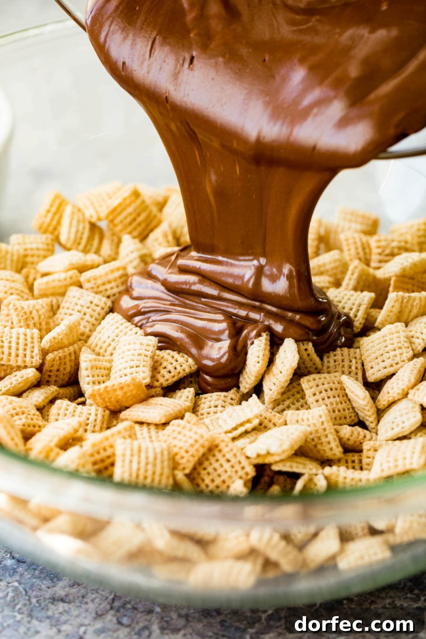 Melted chocolate and peanut butter being poured over rice Chex in a glass bowl.