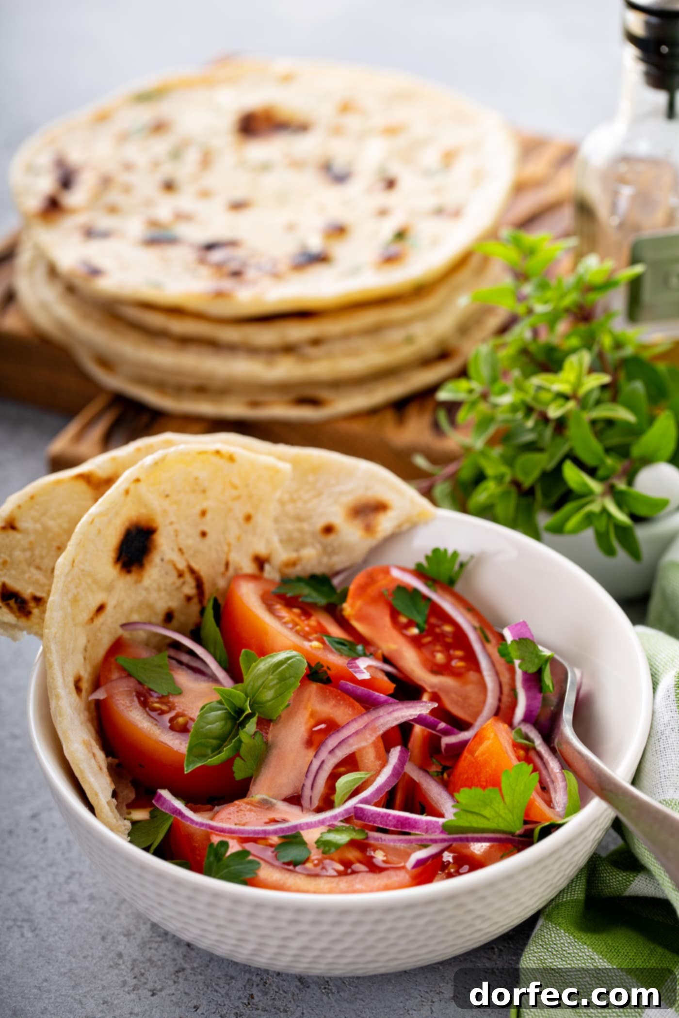 A bowl of fresh tomato and onion salad, with torn pieces of unleavened bread on the side and a stack of bread in the background.