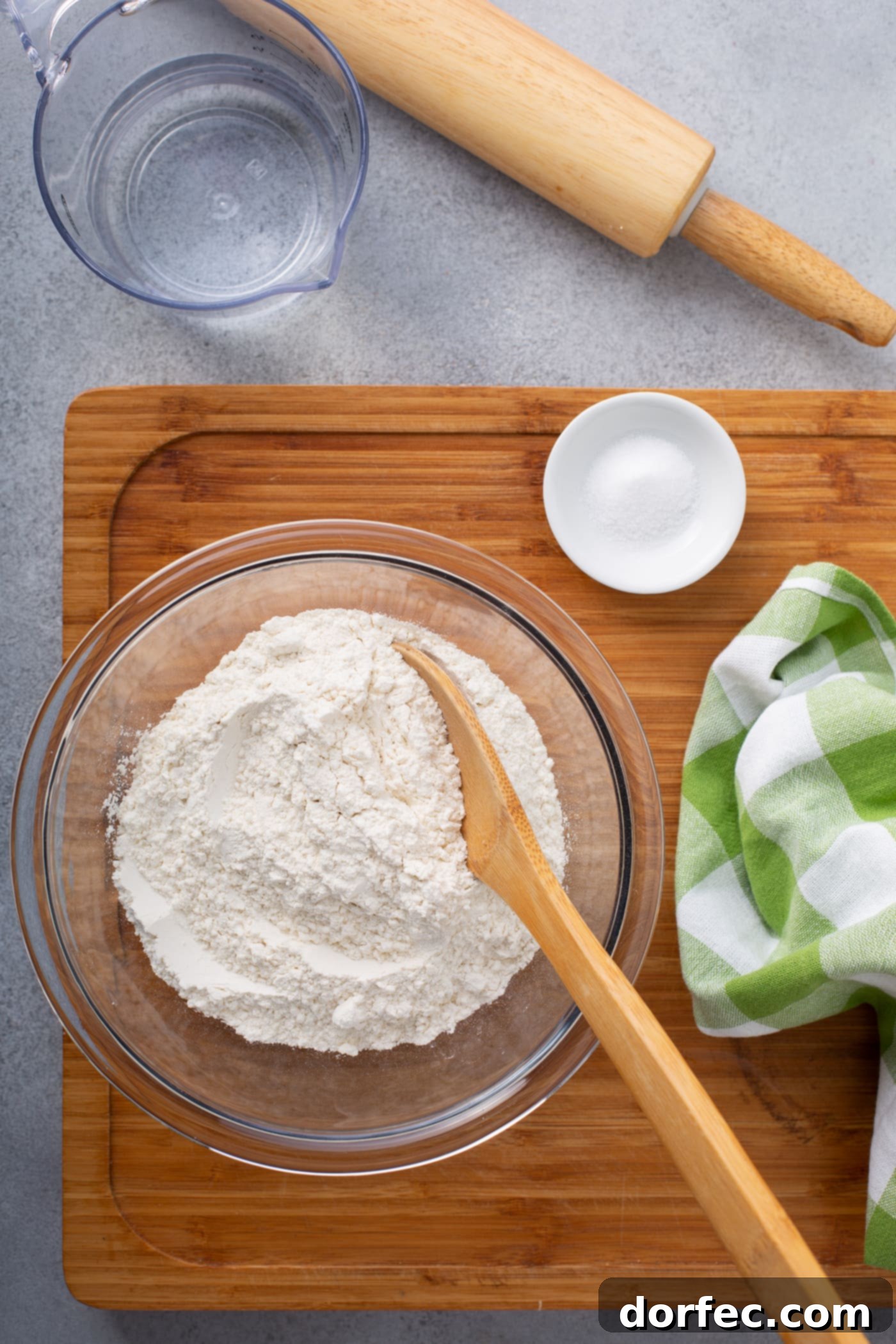 Ingredients for unleavened bread: a glass bowl of flour, a small white bowl of salt, and a glass measuring cup of cold water on a wooden cutting board.
