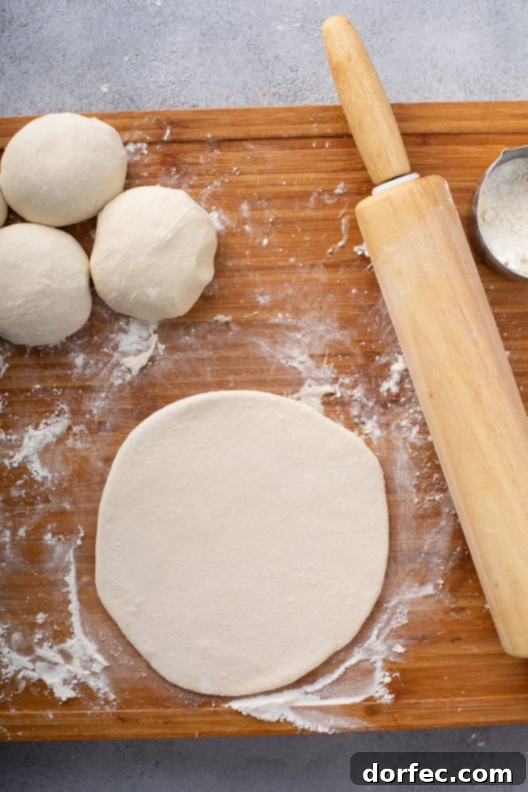 A rolled-out flat circle of unleavened bread dough on a floured wooden board, with other dough portions waiting.