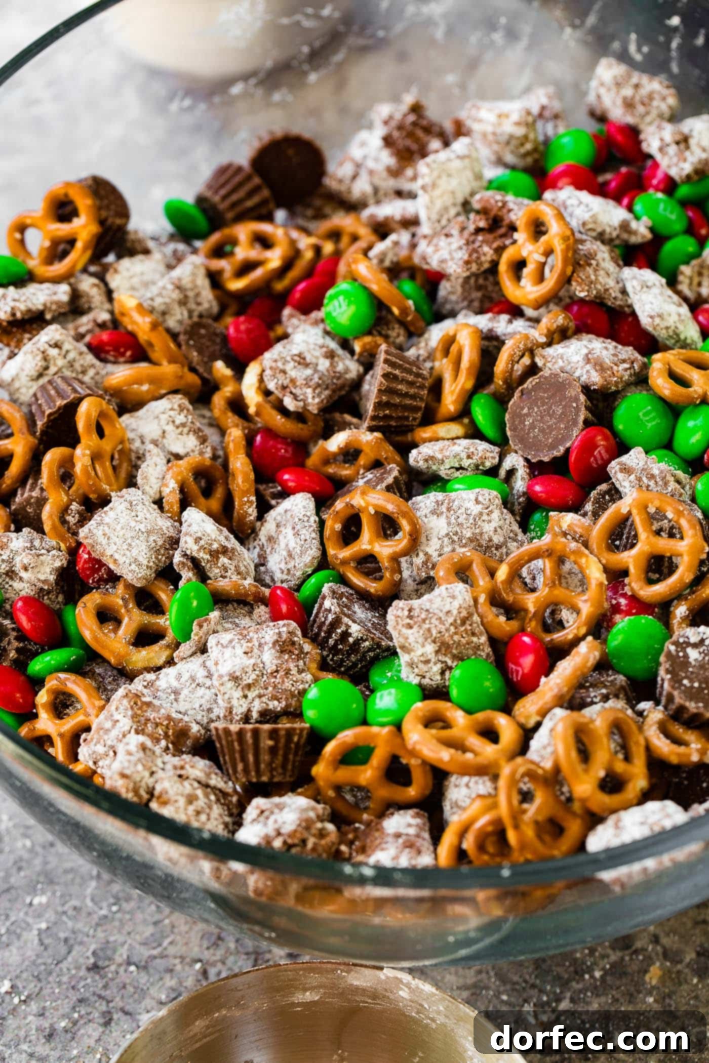 A close-up of a large glass bowl showcasing a delectable Christmas snack mix. The mix is a delightful medley of crunchy pretzels, colorful red and green M&Ms, rich peanut butter cups, and generously coated Chex Muddy Buddies, perfect for holiday indulgence.
