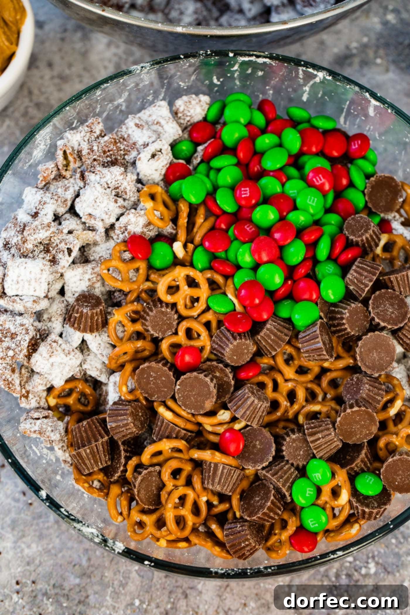 A close-up shot of a large glass bowl filled with a delightful Christmas snack mix, featuring the irresistible combination of sweet and salty pretzels, creamy M&Ms, rich peanut butter cups, and crispy Chex Muddy Buddies.