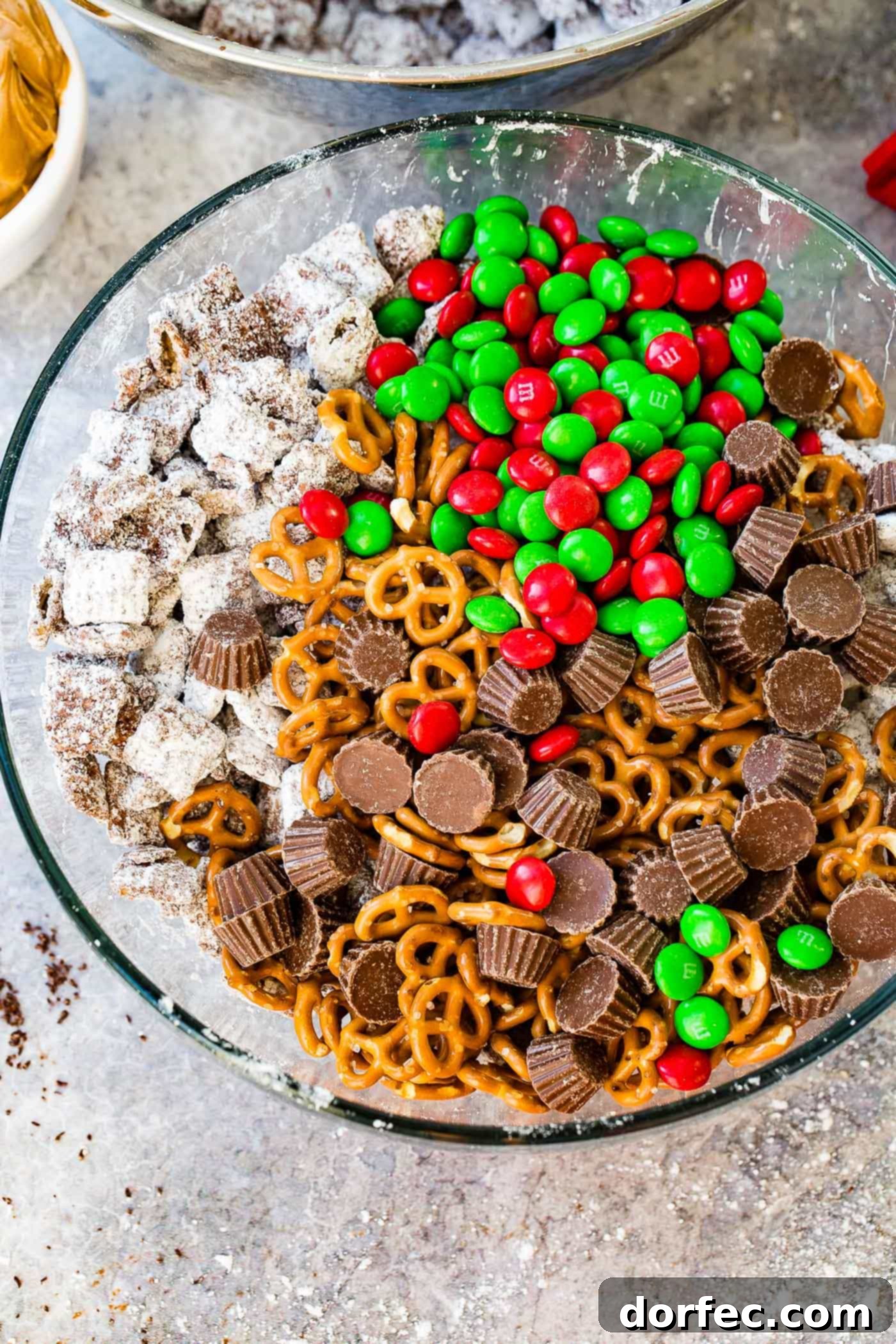 A large glass bowl filled with an irresistible Christmas snack mix, combining the classic flavors of chocolate and peanut butter, highlighted by Chex Muddy Buddies, pretzels, M&Ms, and peanut butter cups.