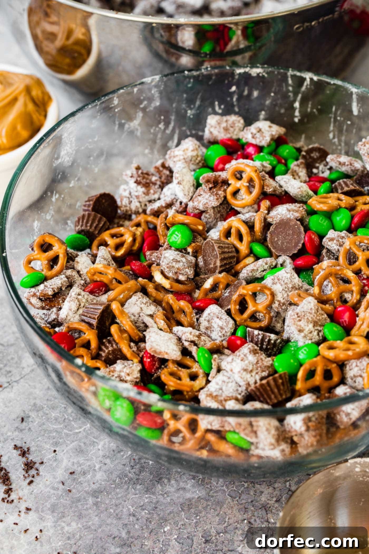 A large glass bowl brimming with a festive Christmas snack mix, featuring a delightful blend of Chex Muddy Buddies, crunchy pretzels, colorful M&Ms, and rich peanut butter cups, perfect for grabbing by the handful.