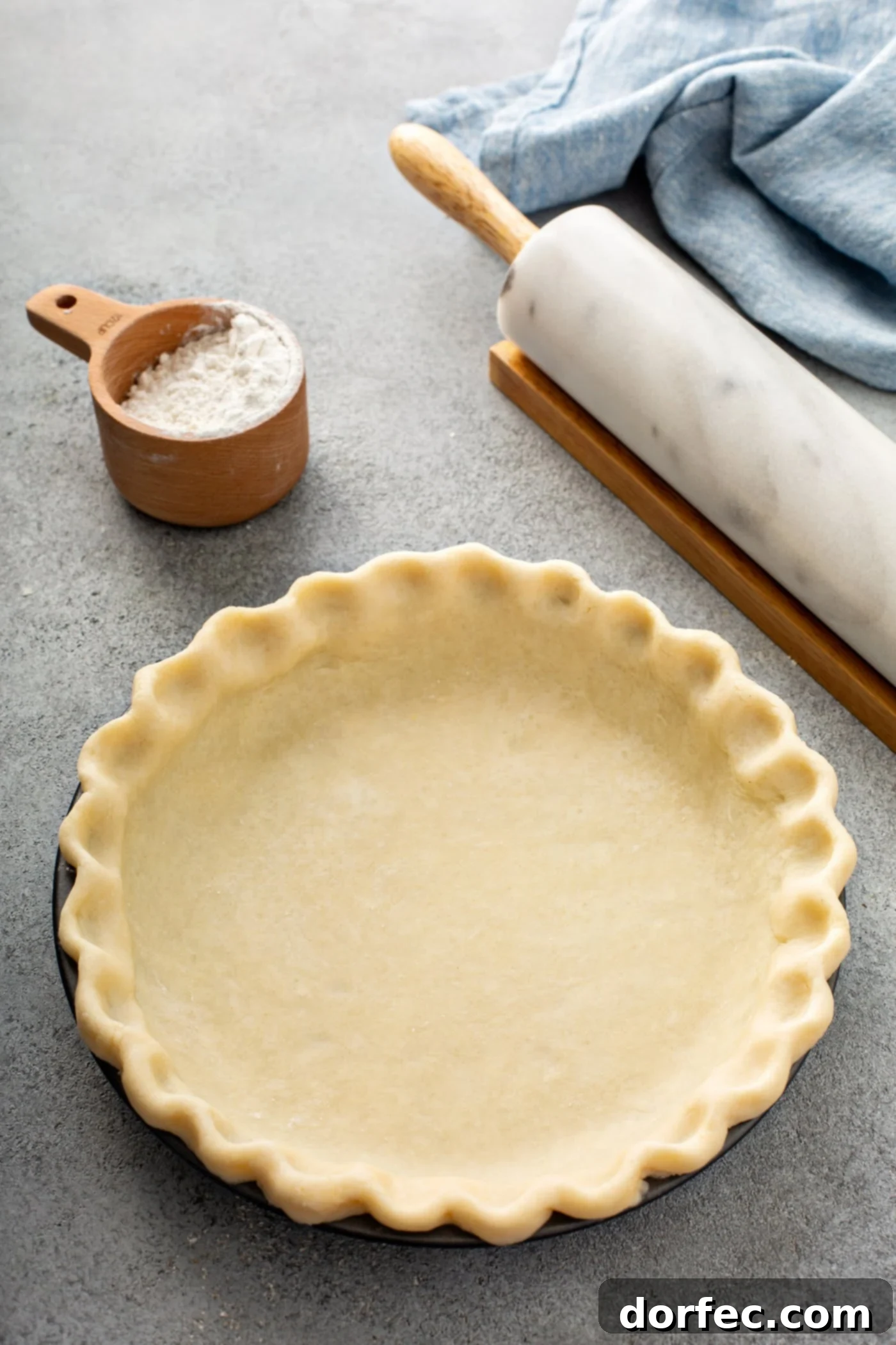 An uncooked all-butter pie crust nestled in a pie tin, ready for filling and baking. A rolling pin and a cup of flour rest nearby on a rustic wooden table.