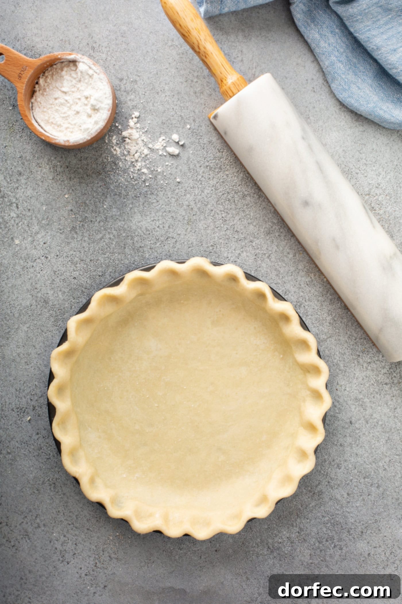 A pristine, uncooked all-butter pie crust neatly crimped and ready in a pie tin, sitting on a table alongside a rolling pin and a cup of flour, awaiting its delicious fate.