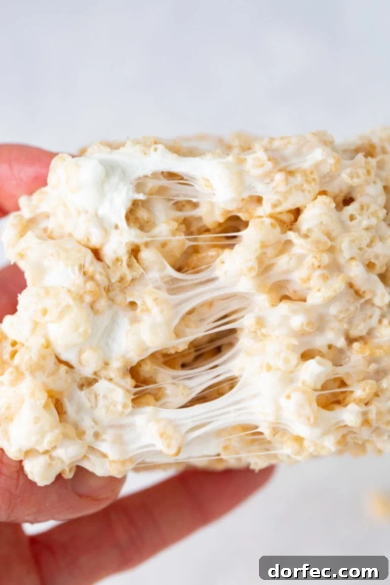 Extra Gooey Rice Krispie Treats 3 A perfectly gooey piece of Rice Krispie Treat being gently pulled apart, revealing the luscious melted marshmallow stretching between the crispy cereal grains, a testament to its soft and chewy texture.