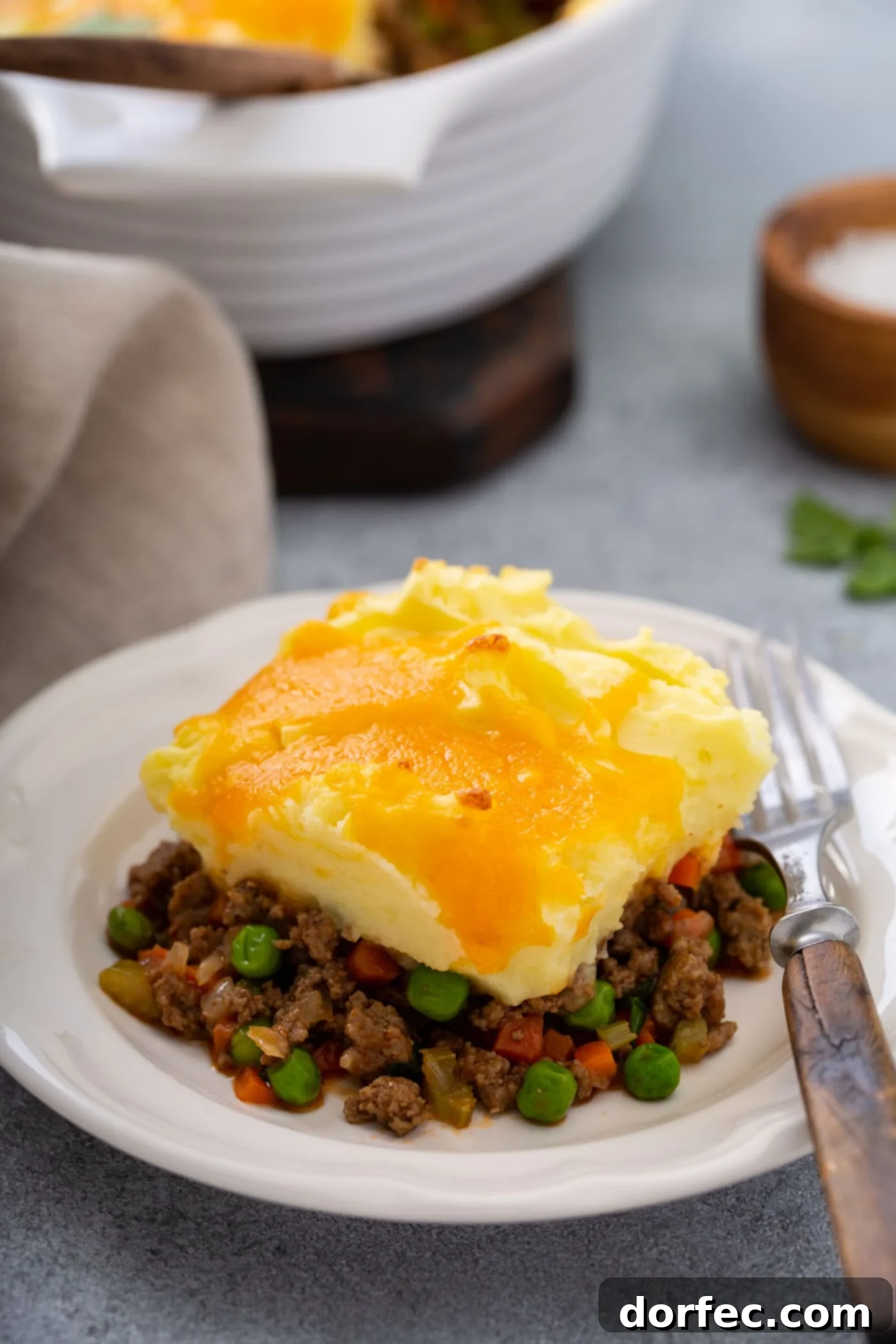 A serving of shepherd's pie on a white plate with a fork beside it. There is a napkin and a casserole dish in the background.
