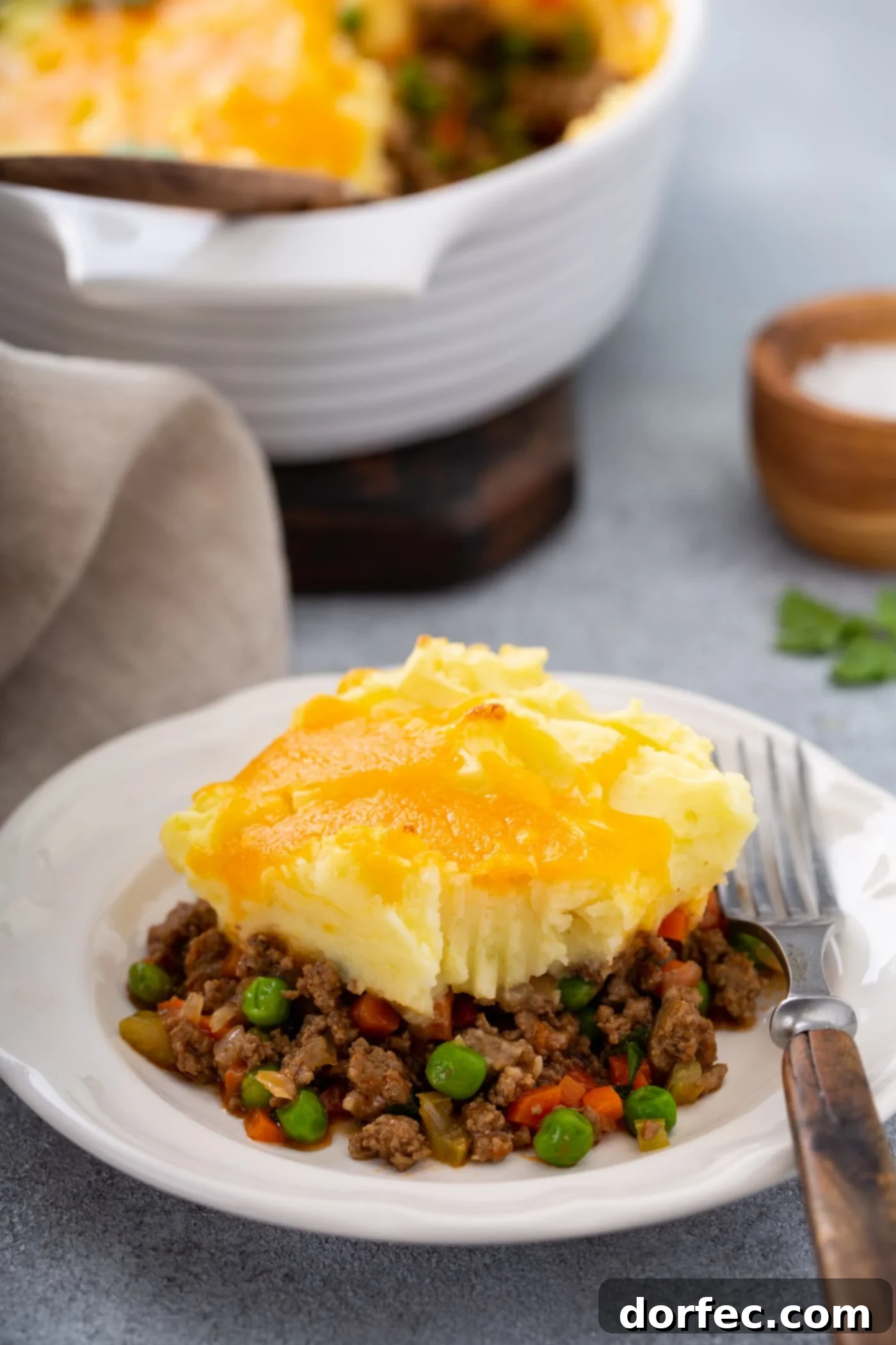 A serving of shepherd's pie with carrots, peas and ground beef for filling, topped with mashed potatoes and melted cheese. There is a fork resting on the plate and a napkin and a small container of salt next to it.