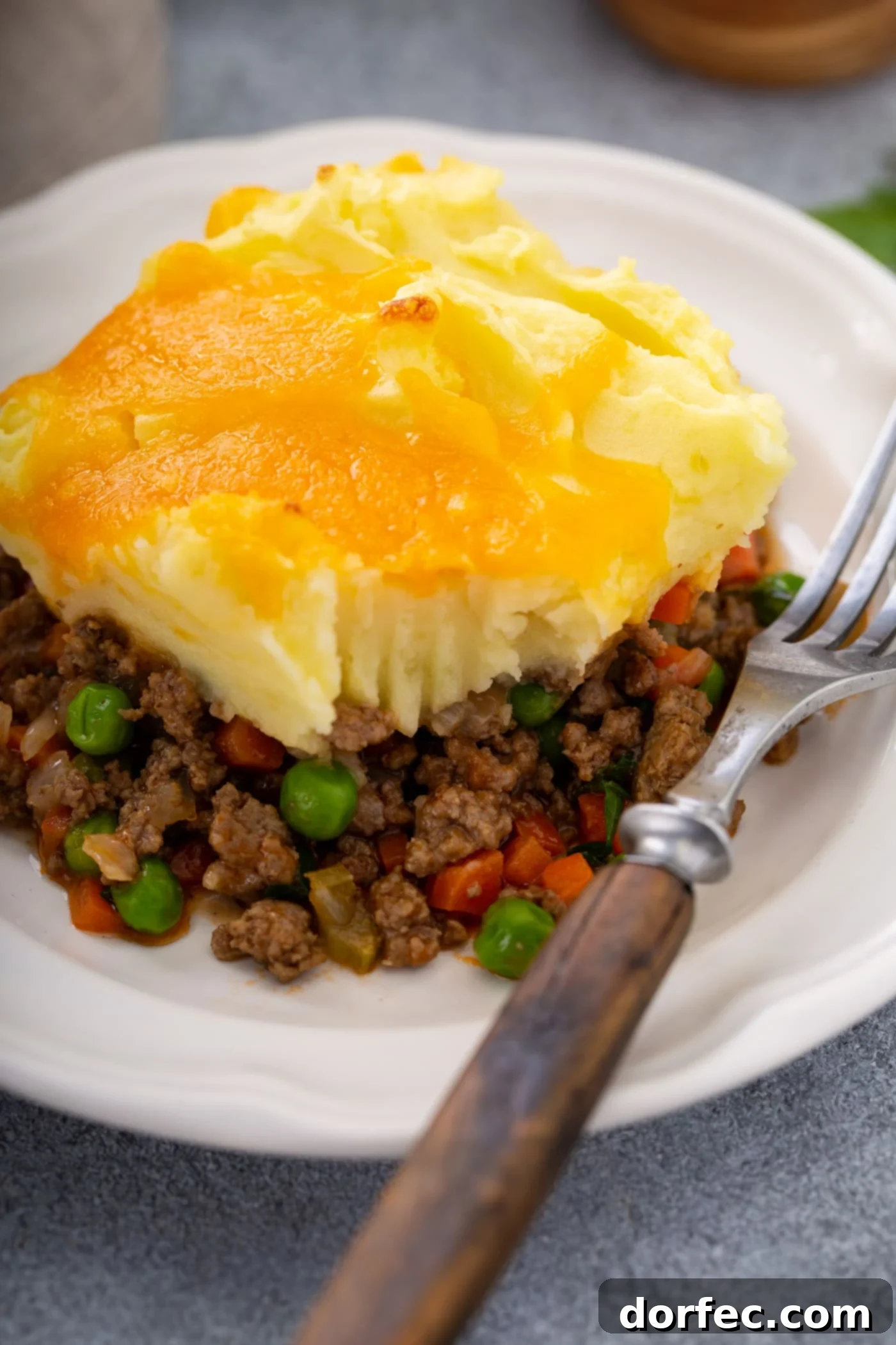 A serving of shepherd's pie on a white plate with a fork resting beside it. You can see the rich ground beef and vegetable filling, the mashed potatoes on top, and the yellow melted cheddar cheese topping it all.