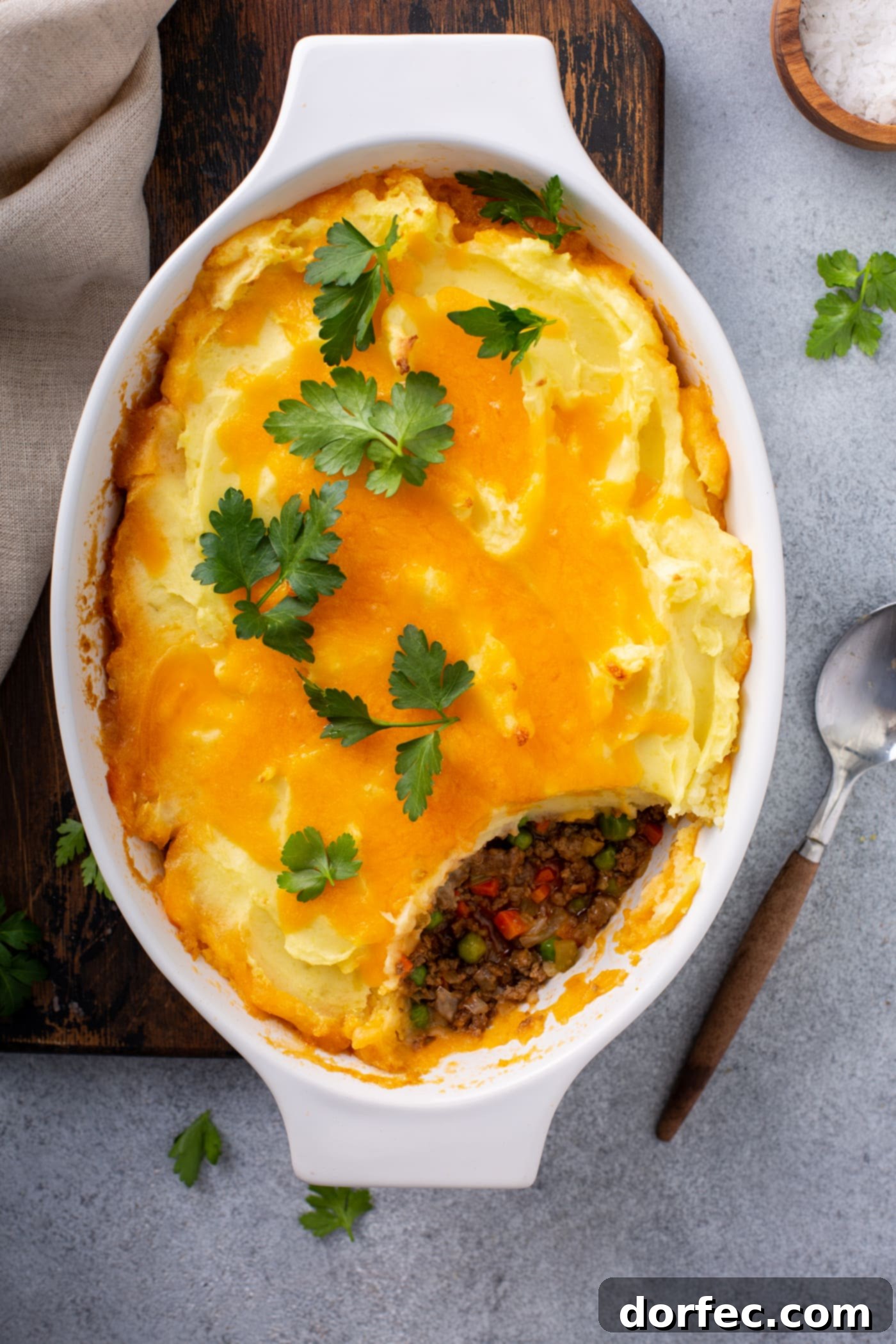 A casserole dish of shepherd's pie. A serving has been removed so you can see the ground beef, carrots and peas in the filling. It is topped with mashed potatoes and melted cheese with a few parsley leaves on top.