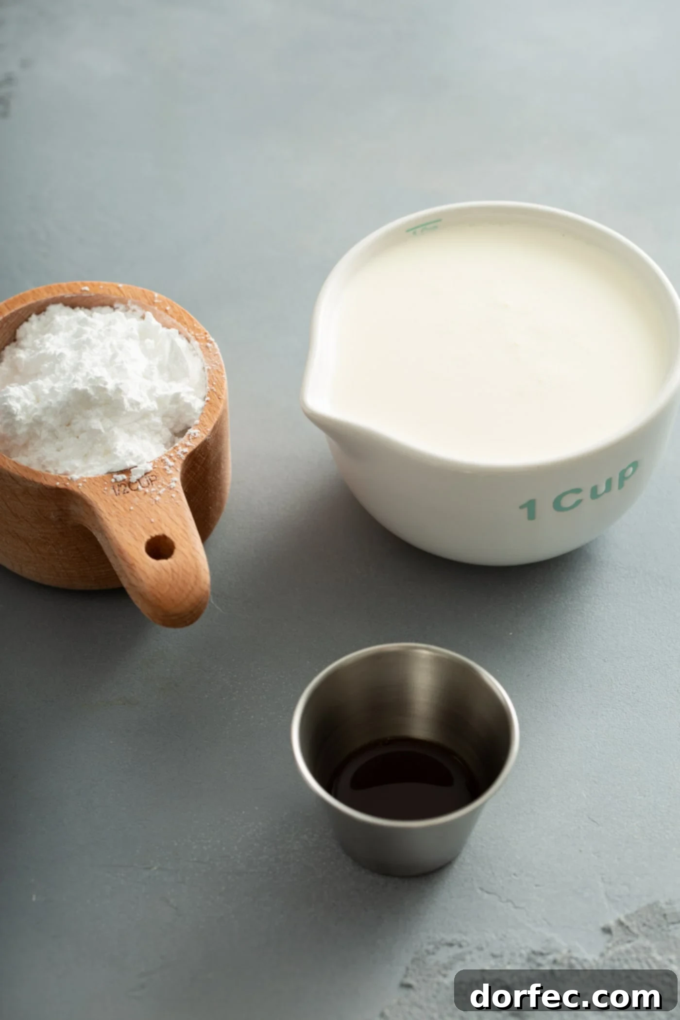 A clear measuring cup filled with heavy cream, a smaller measuring cup containing fine powdered sugar, and a petite silver container of vanilla extract are neatly arranged on a surface, showcasing the three simple ingredients required for homemade whipped cream.