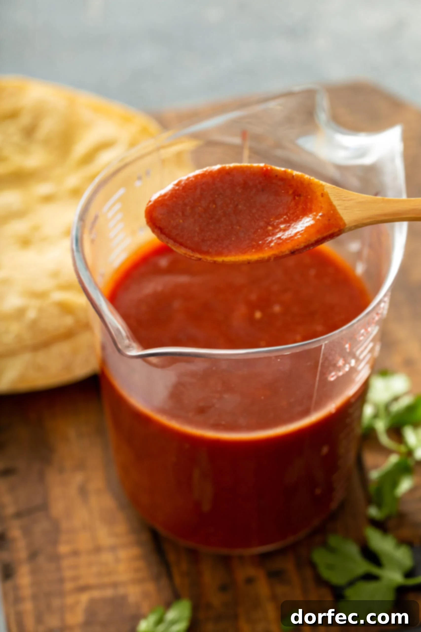 A clear glass measuring cup holds a generous amount of rich, homemade red enchilada sauce, with a wooden spoon dipped into its smooth texture. A blurred background of tortillas and fresh cilantro adds to the inviting culinary scene.
