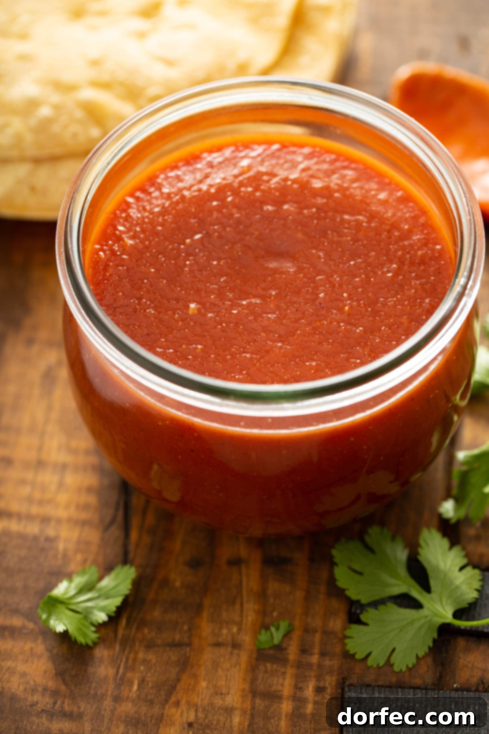 A beautiful glass jar brimming with homemade red enchilada sauce. In the background, artfully arranged tortillas, fresh cilantro, and ripe tomatoes create an inviting culinary tableau.