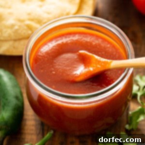 A glass container with a wooden spoon. The container is filled with red enchilada sauce. There are tortillas, green chilies, and cilantro leaves surrounding the container of red sauce.