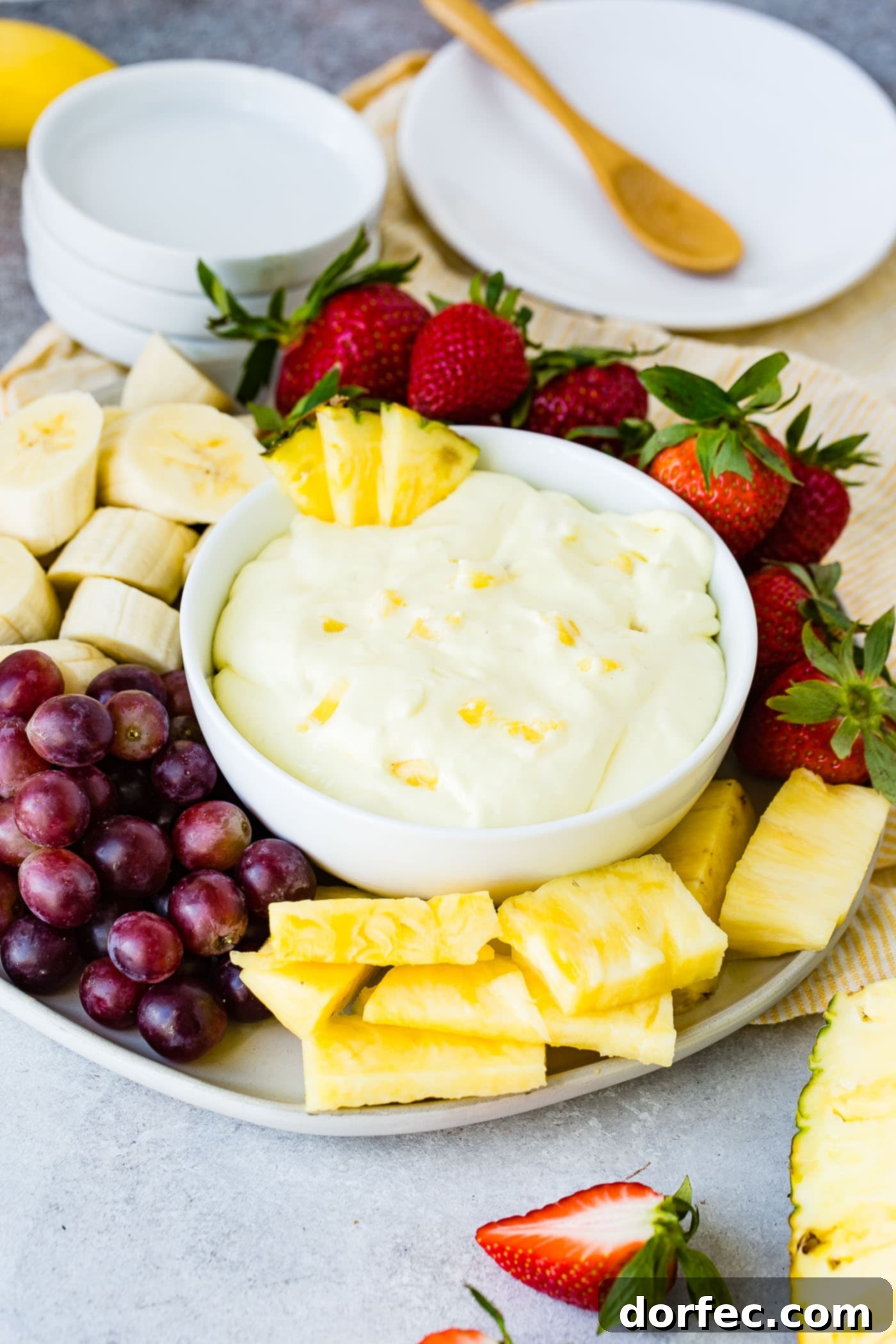 A vibrant bowl of pineapple cream cheese fruit dip, garnished with three fresh pineapple chunks on the rim. The bowl rests on an elegant serving tray, surrounded by a colorful assortment of sliced bananas, plump red grapes, additional pineapple pieces, and juicy strawberries, creating an inviting display.