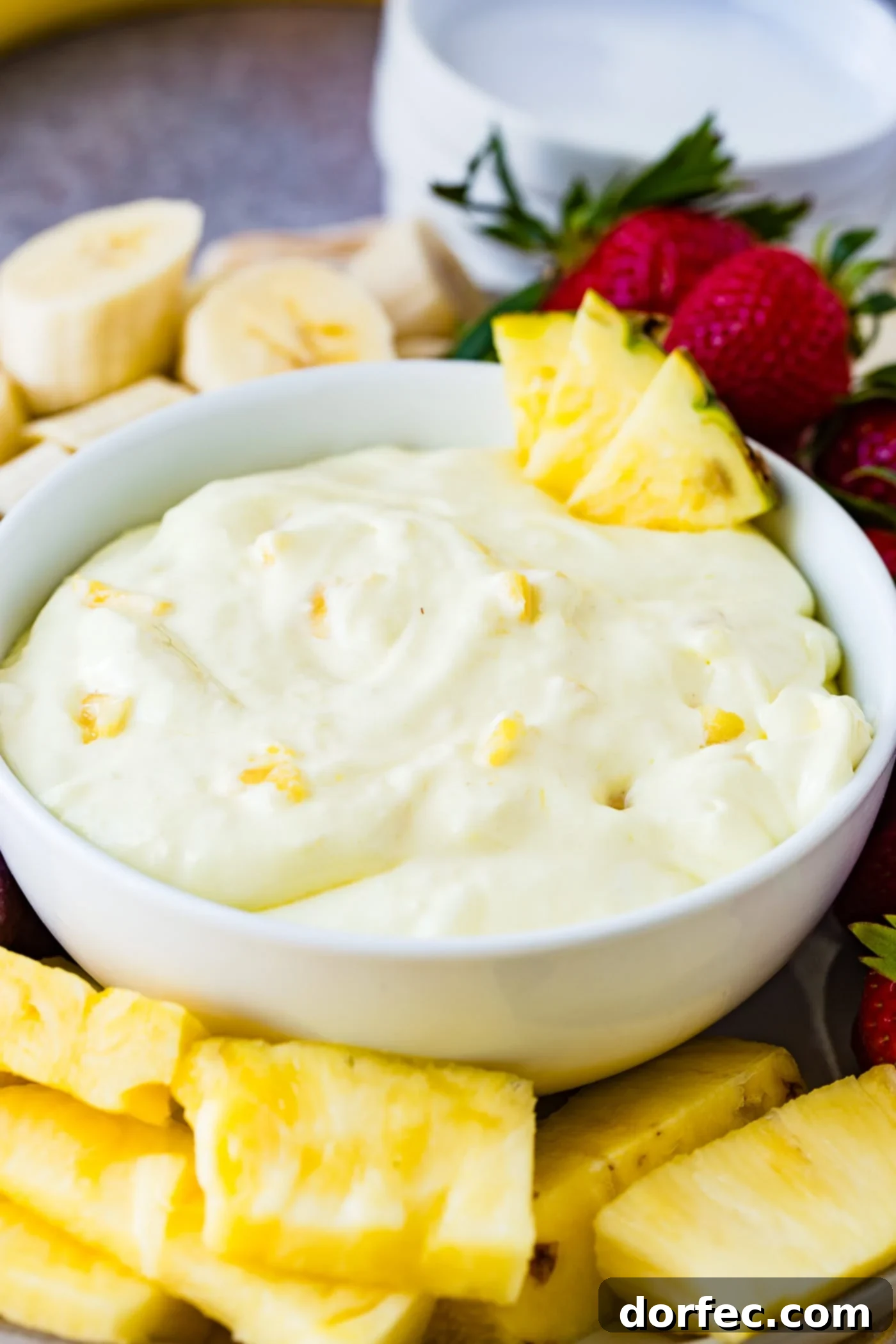 A pristine white bowl filled with creamy pineapple cream cheese fruit dip. Four golden pineapple chunks adorn the edge, with delicate bits of pineapple visible throughout the dip. The bowl is beautifully arranged on a platter, encircled by perfectly sliced bananas, whole ripe strawberries, glistening red grapes, and more succulent pineapple pieces, creating a tempting and colorful fruit spread.
