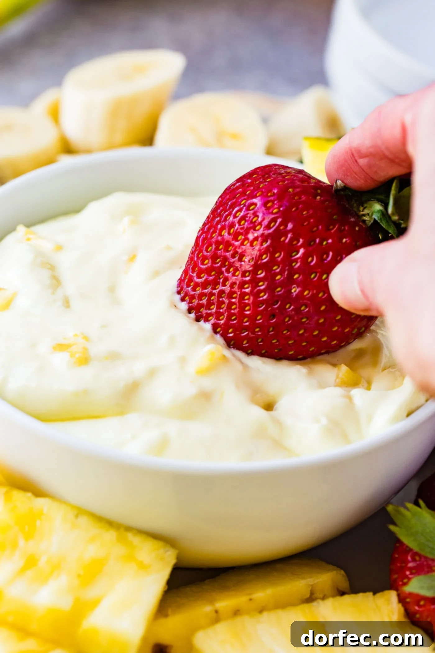 A vibrant red strawberry poised mid-dip into a pristine white bowl of pineapple fruit dip, with tiny, delectable pieces of pineapple visible within the creamy texture. The bowl of dip is artfully surrounded by an appetizing array of perfectly sliced bananas and succulent pineapple chunks, inviting a taste of this tropical treat.