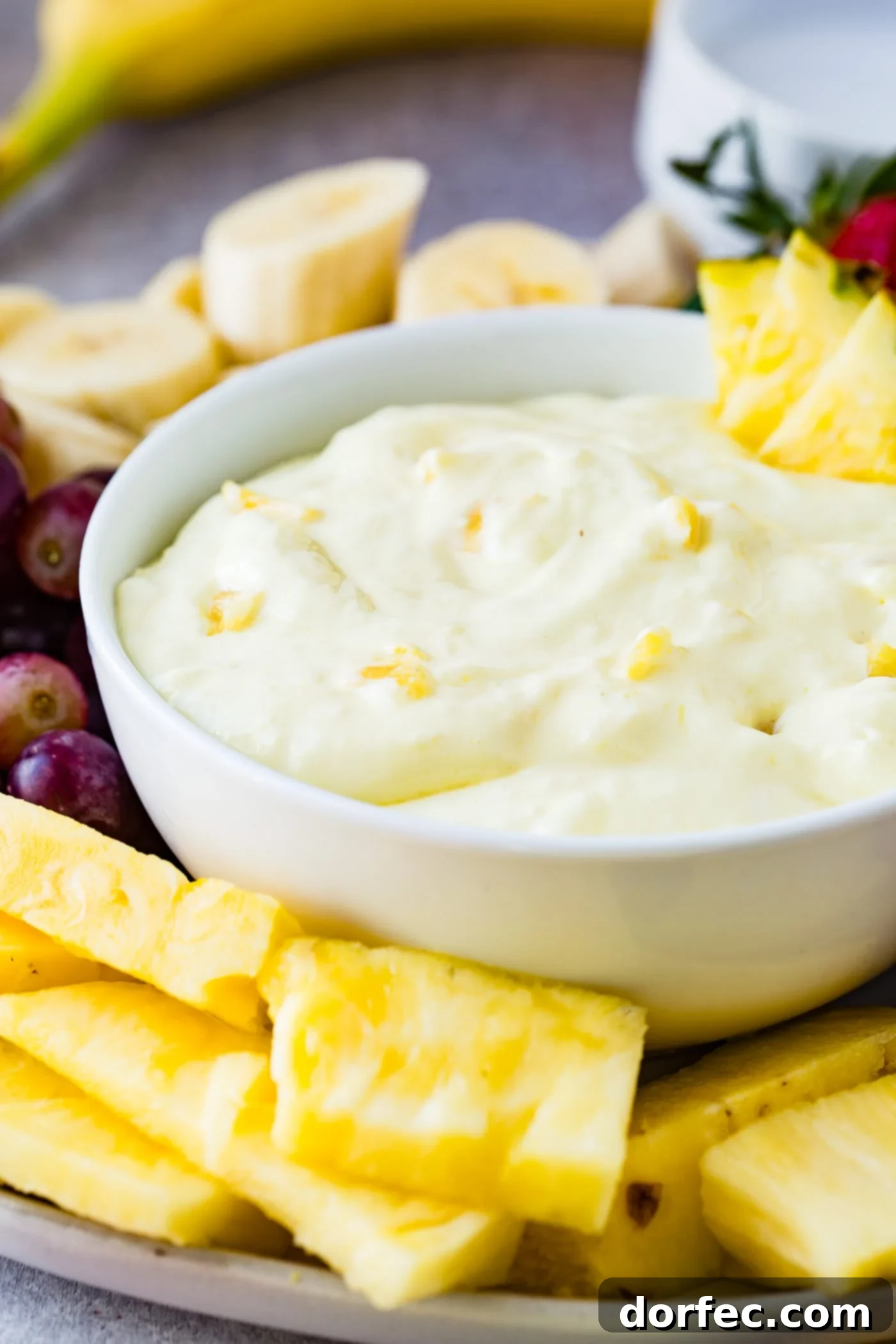 A pristine white bowl showcasing the pineapple fruit dip, with three vibrant pineapple chunks resting elegantly on its rim and delicate pieces of pineapple mixed throughout the creamy texture. The bowl is artfully surrounded by an appetizing display of perfectly sliced bananas, sweet red grapes, more succulent pineapple chunks, and whole luscious strawberries, presenting a delightful array for dipping.