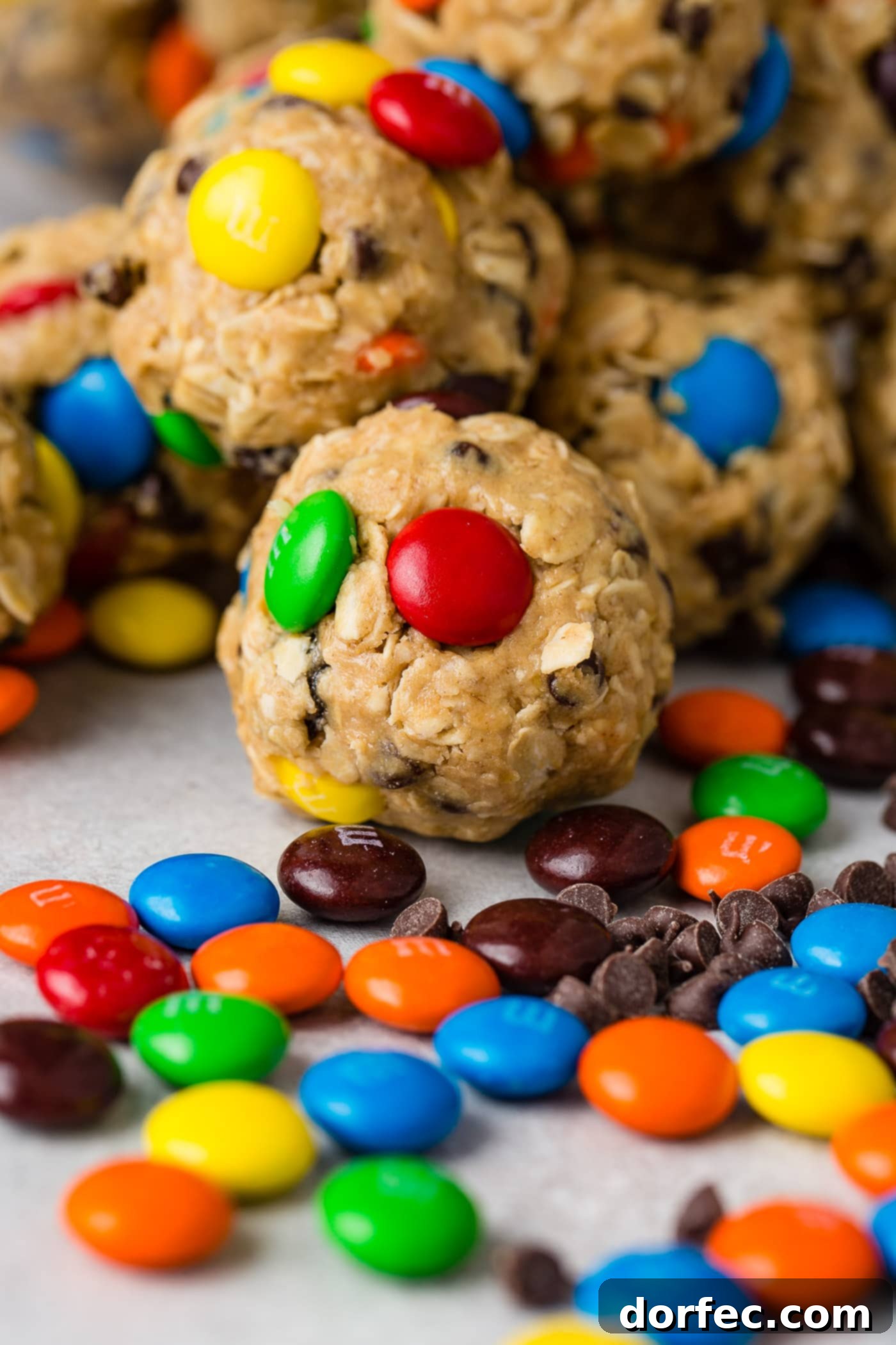 Protein-Packed Peanut Butter Monster Cookie Energy Bites 4 A close-up shot of seven perfectly formed no-bake protein monster energy balls, laden with oats, raisins, mini M&M's, and mini chocolate chips. A scattering of mini M&M's and raisins lies on the wooden surface in front, emphasizing their delightful inclusions.