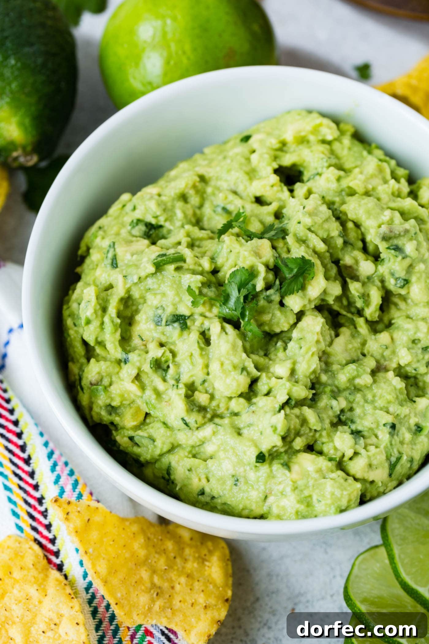 A beautifully presented white bowl of fresh guacamole, garnished with a few cilantro leaves. Nearby, a scattering of golden tortilla chips and slices of fresh lime invite you to dig in.